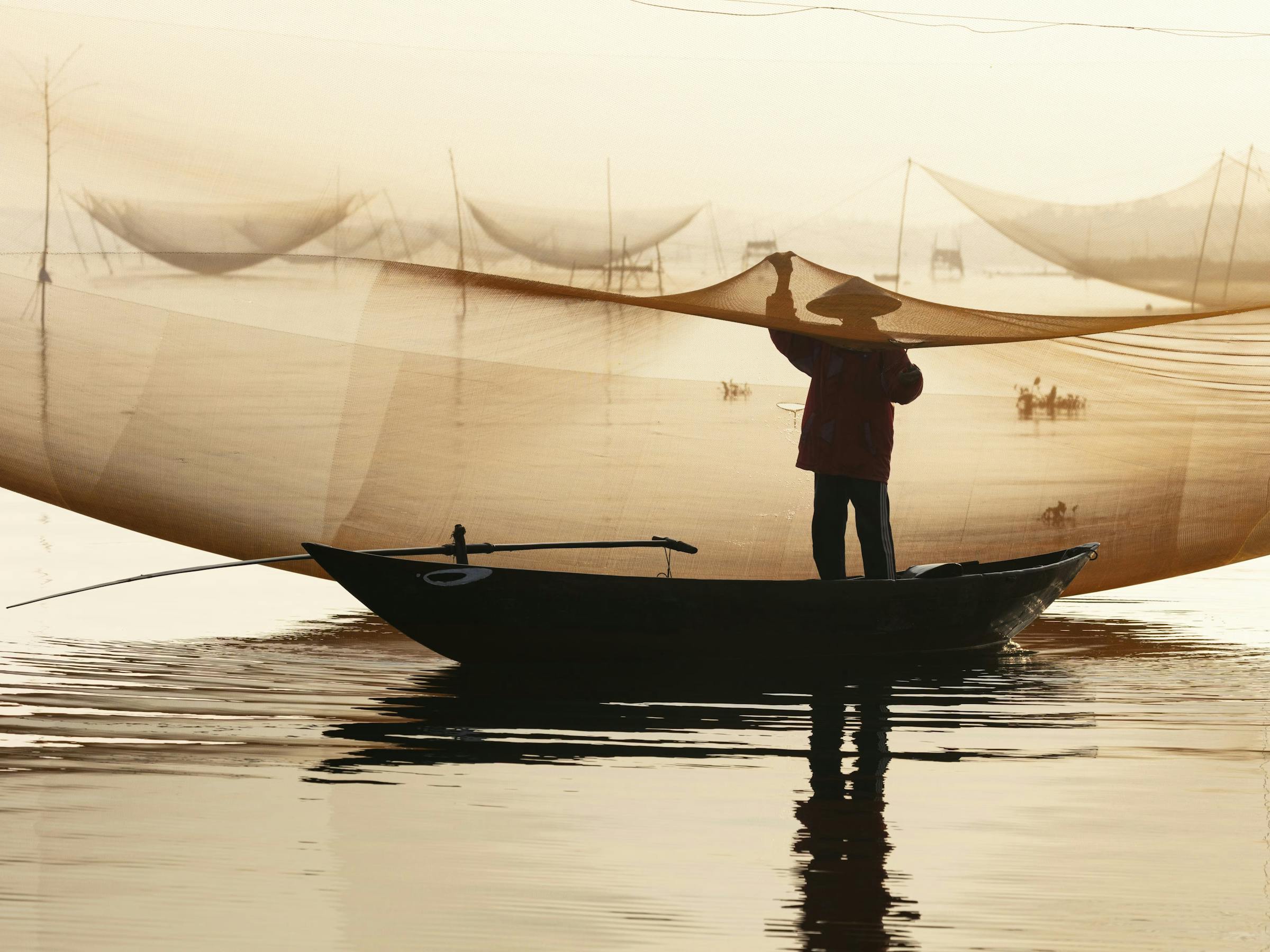 Person on a boat holds fishing nets over calm water at sunrise.