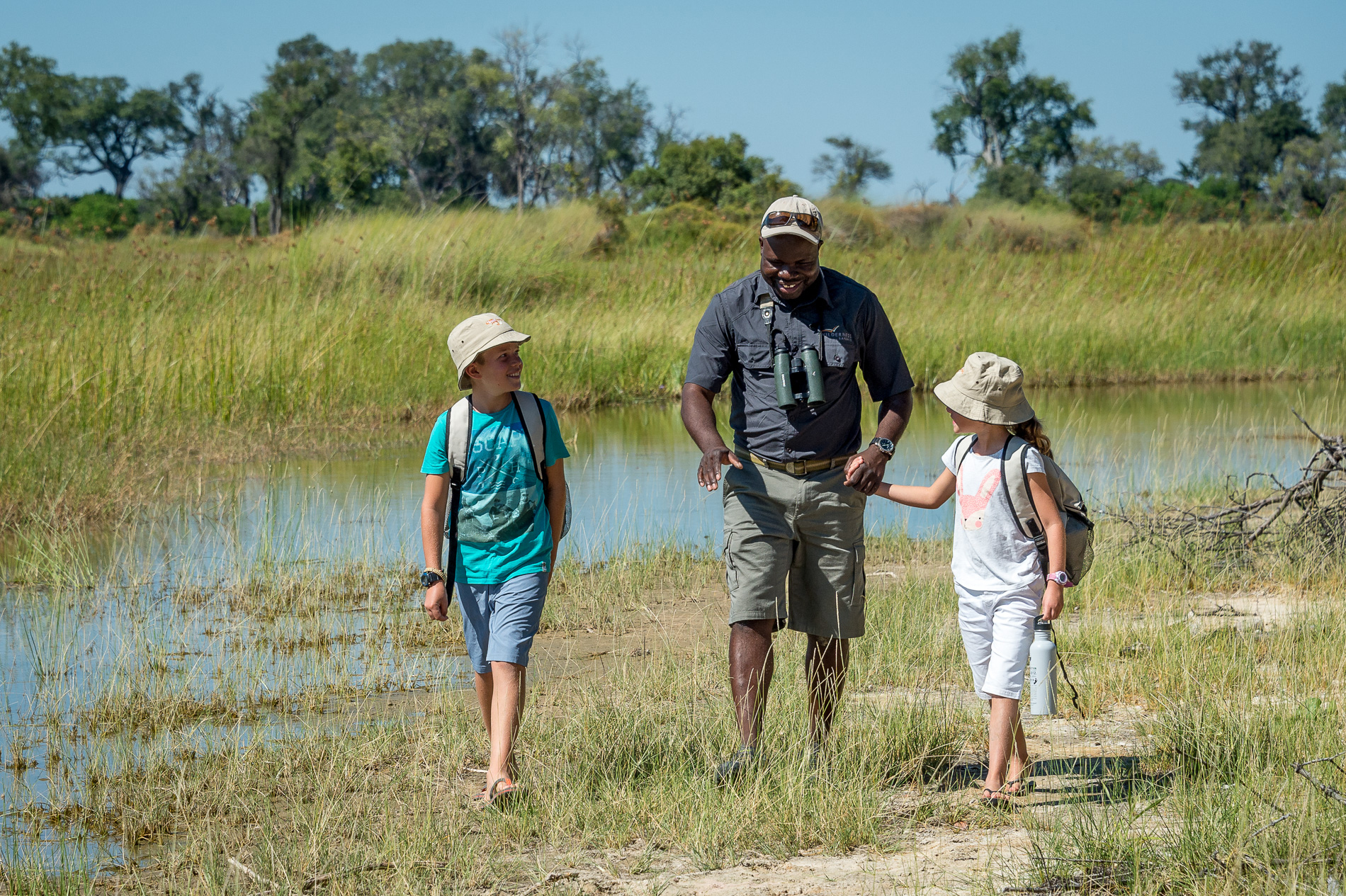 Two children walk beside an adult through shallow floodplain water, with green reeds and trees around them.