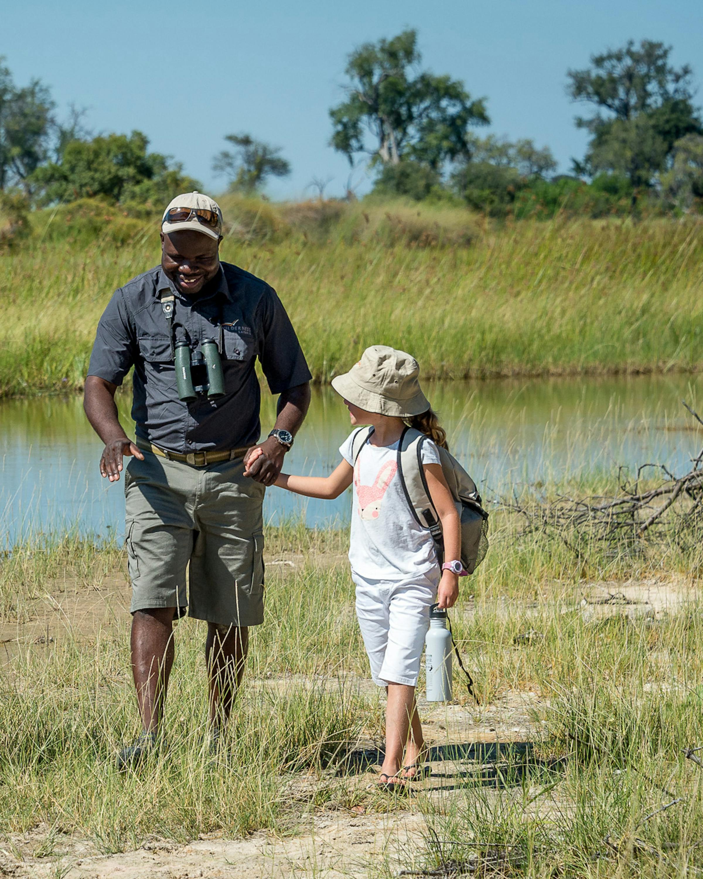 Two children walk beside an adult through shallow floodplain water, with green reeds and trees around them.