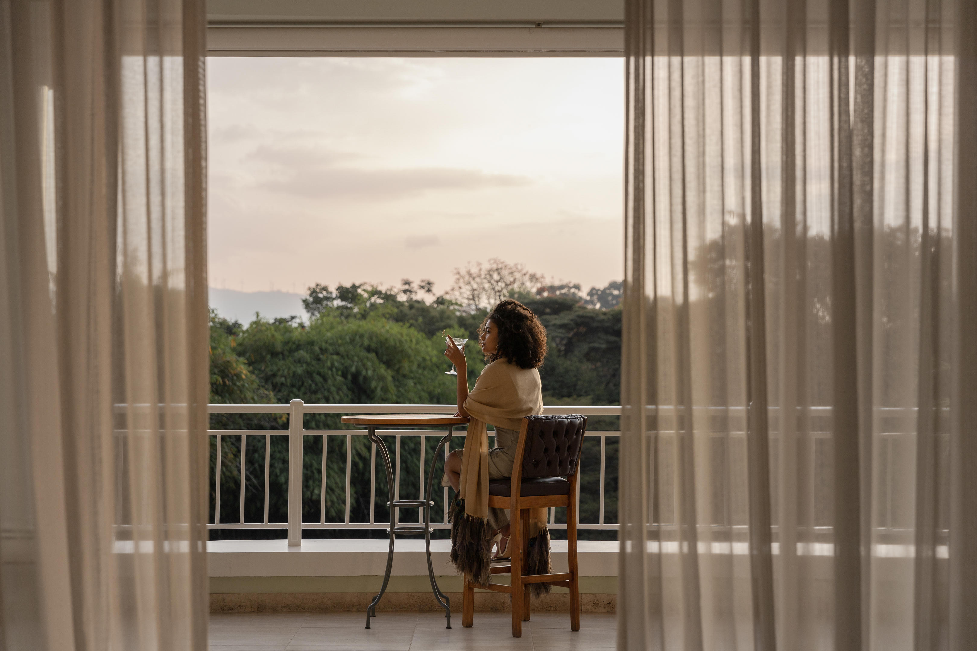 A guest sits on a balcony at Hemingways Nairobi, looking out over treetops through sheer curtains at dusk.