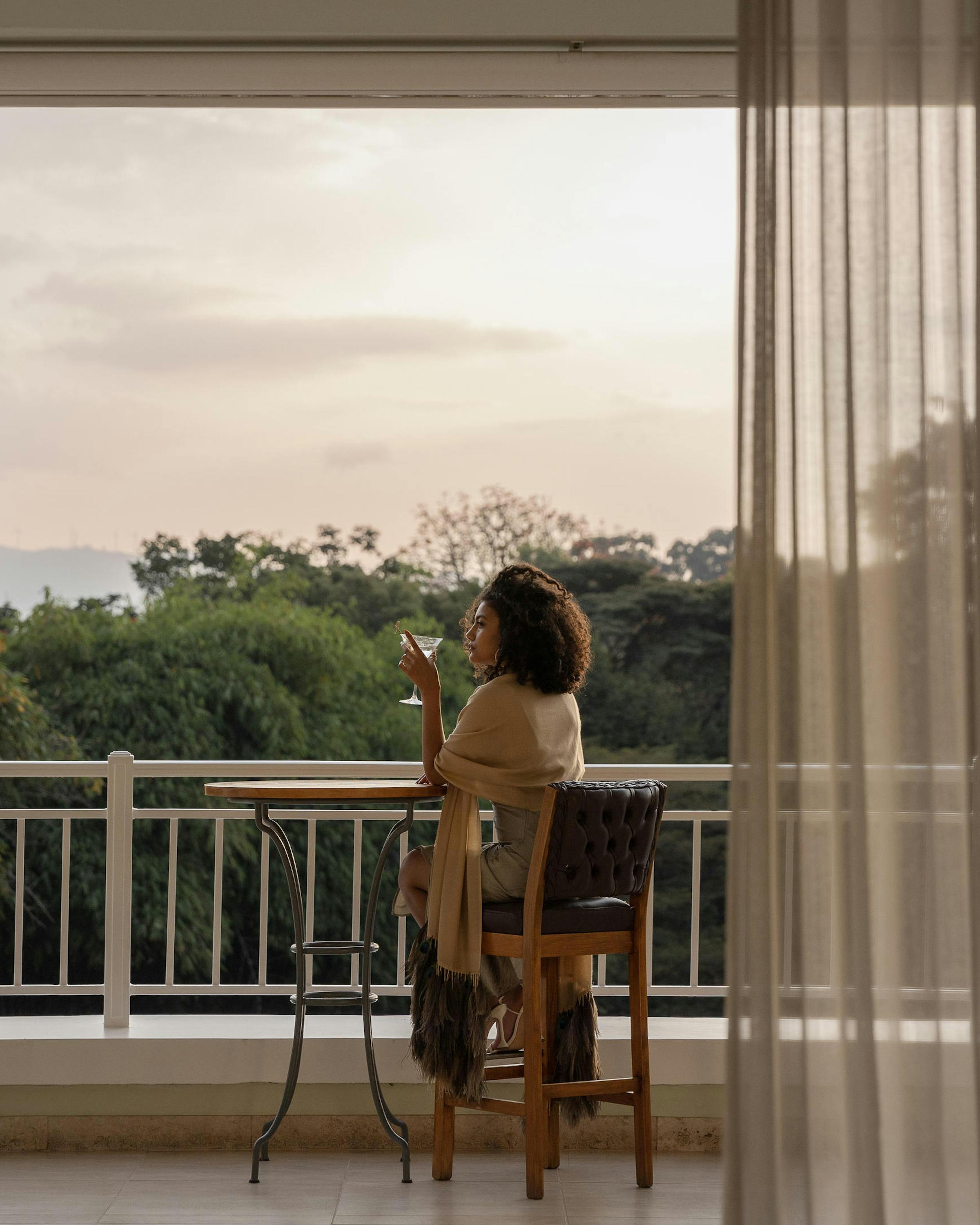 A guest sits on a balcony at Hemingways Nairobi, looking out over treetops through sheer curtains at dusk.