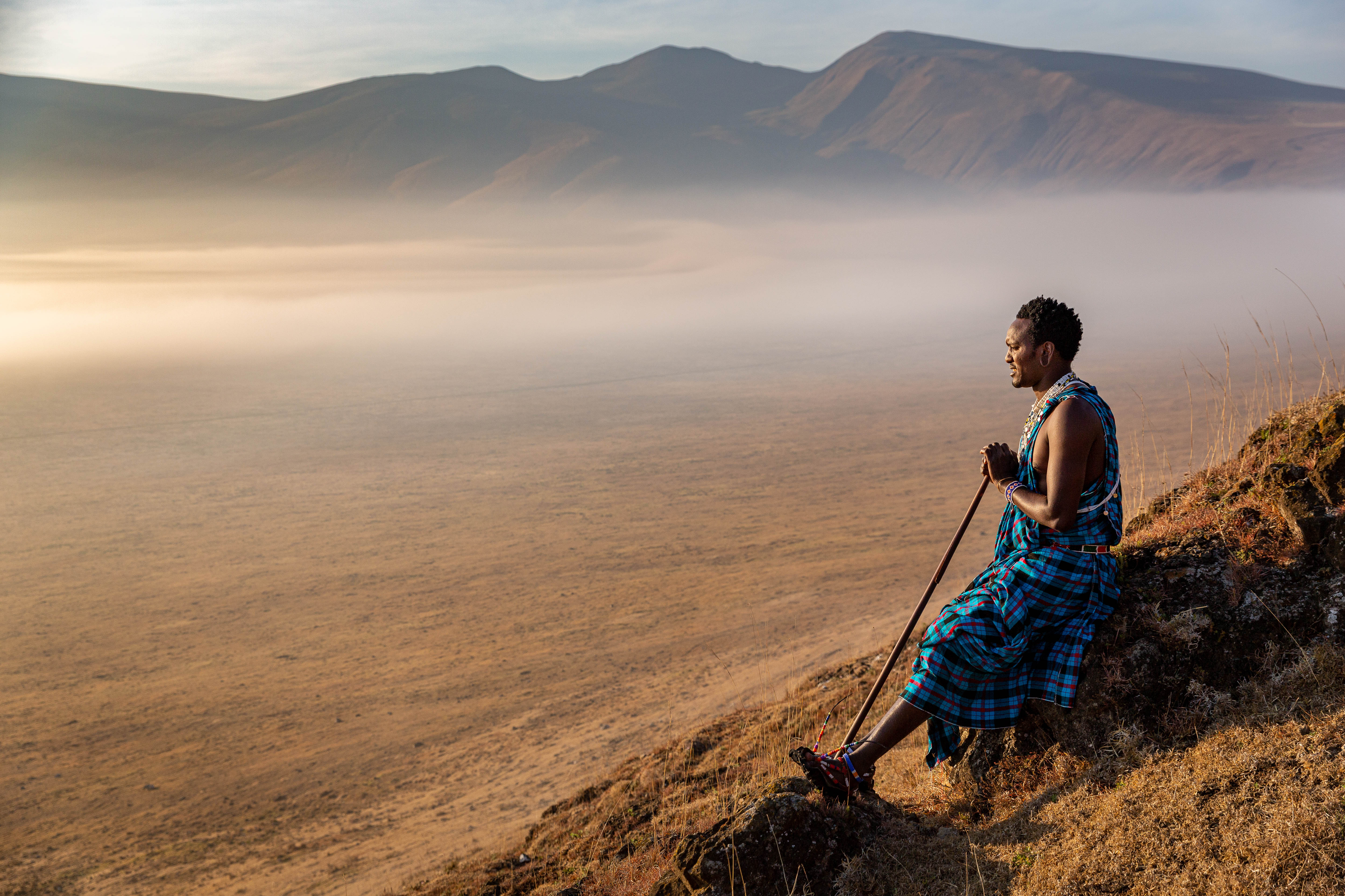 A Maasai person stands on a rocky rim looking over the Ngorongoro Crater, with mist and mountains beyond.