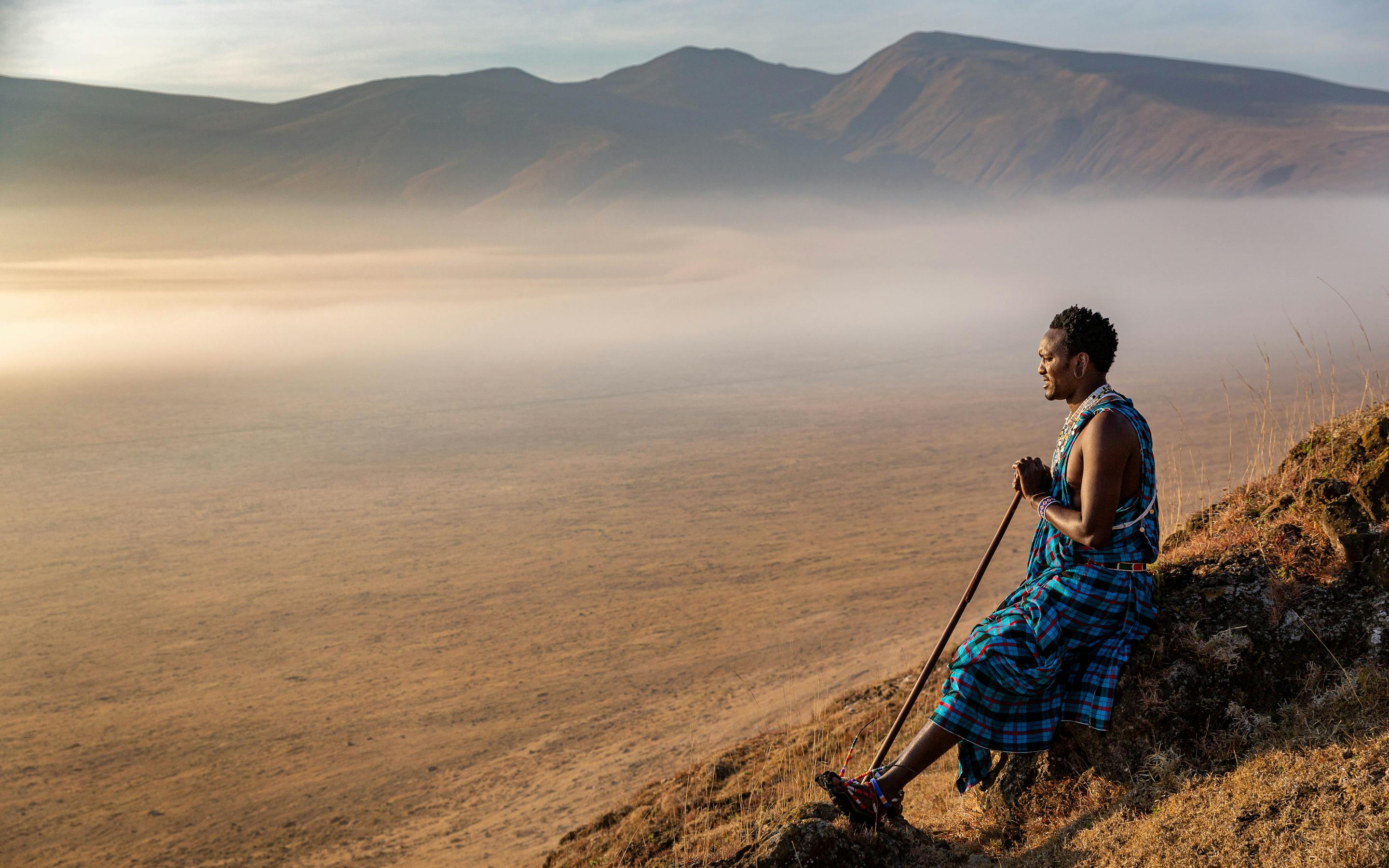 A Maasai person stands on a rocky rim looking over the Ngorongoro Crater, with mist and mountains beyond.