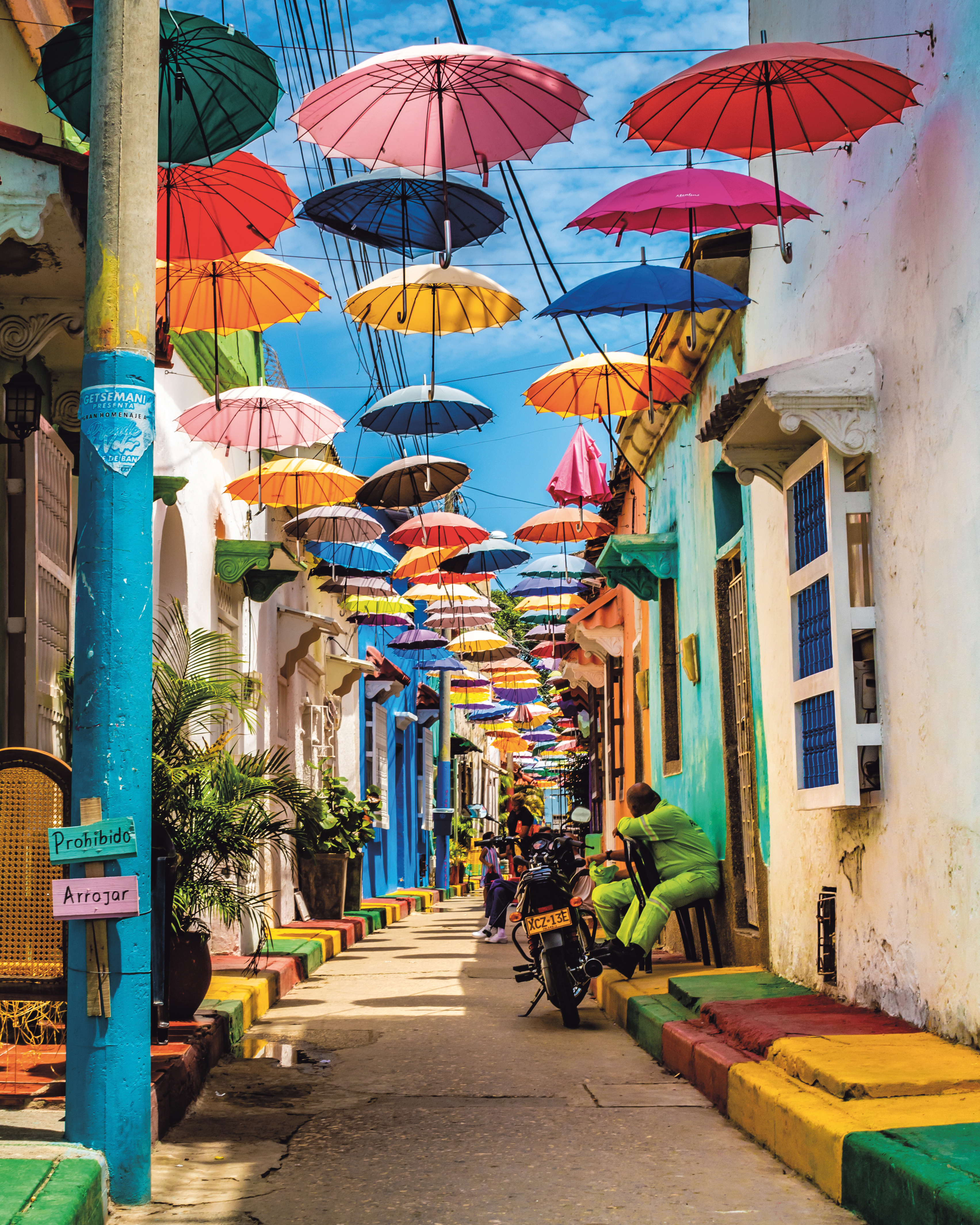A street in Cartagena is strung with colorful umbrellas overhead, casting shade on bright facades and pedestrians.