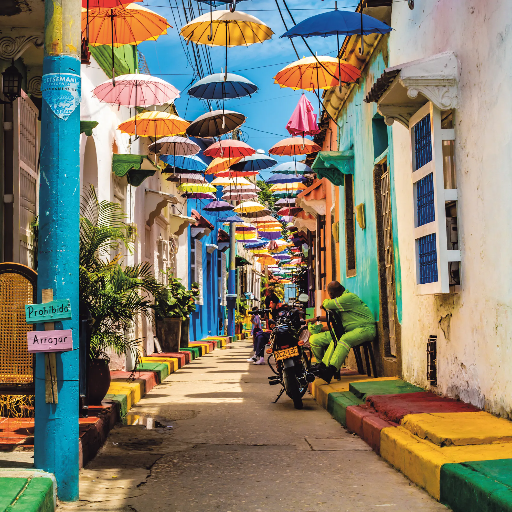 A street in Cartagena is strung with colorful umbrellas overhead, casting shade on bright facades and pedestrians.