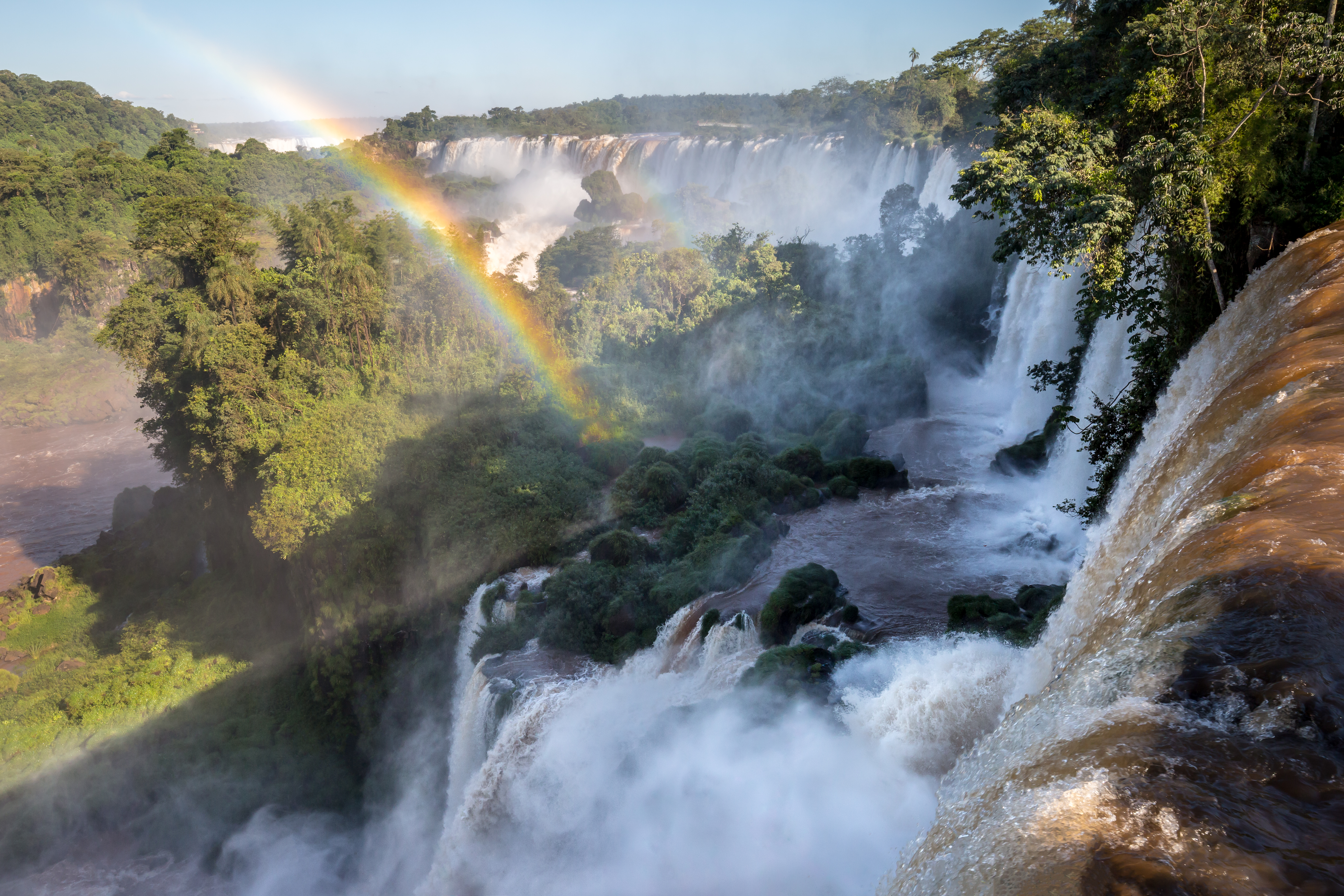 A bright rainbow arcs through mist above Iguazu Falls, with foaming cascades dropping into a forested gorge.