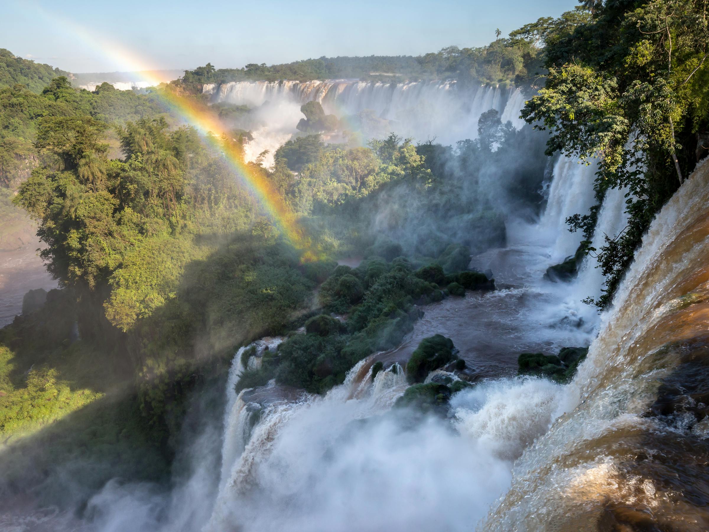A bright rainbow arcs through mist above Iguazu Falls, with foaming cascades dropping into a forested gorge.