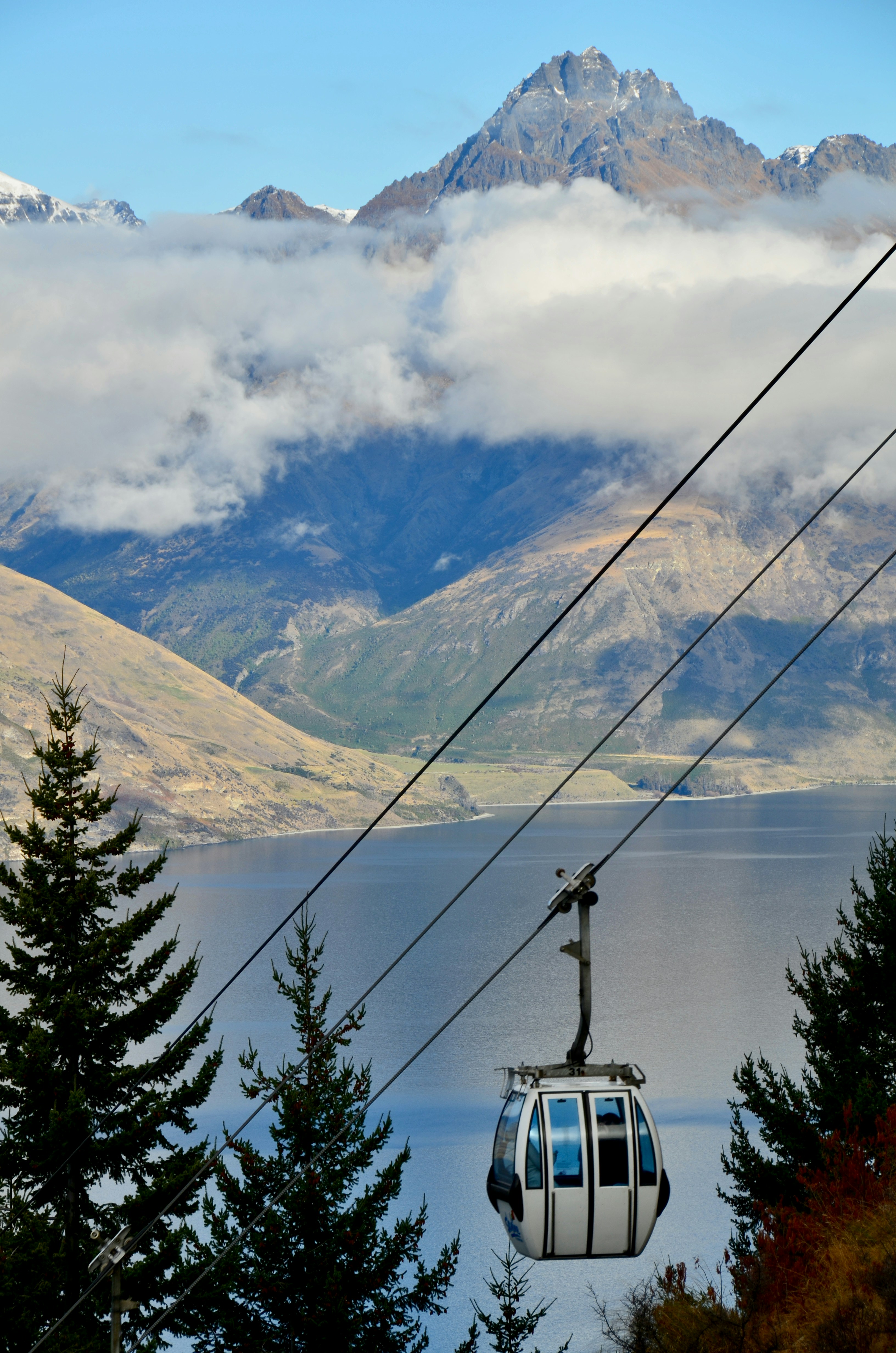 A gondola cabin rides above Queenstown with Lake Wakatipu and mountains behind, low clouds drifting across peaks.