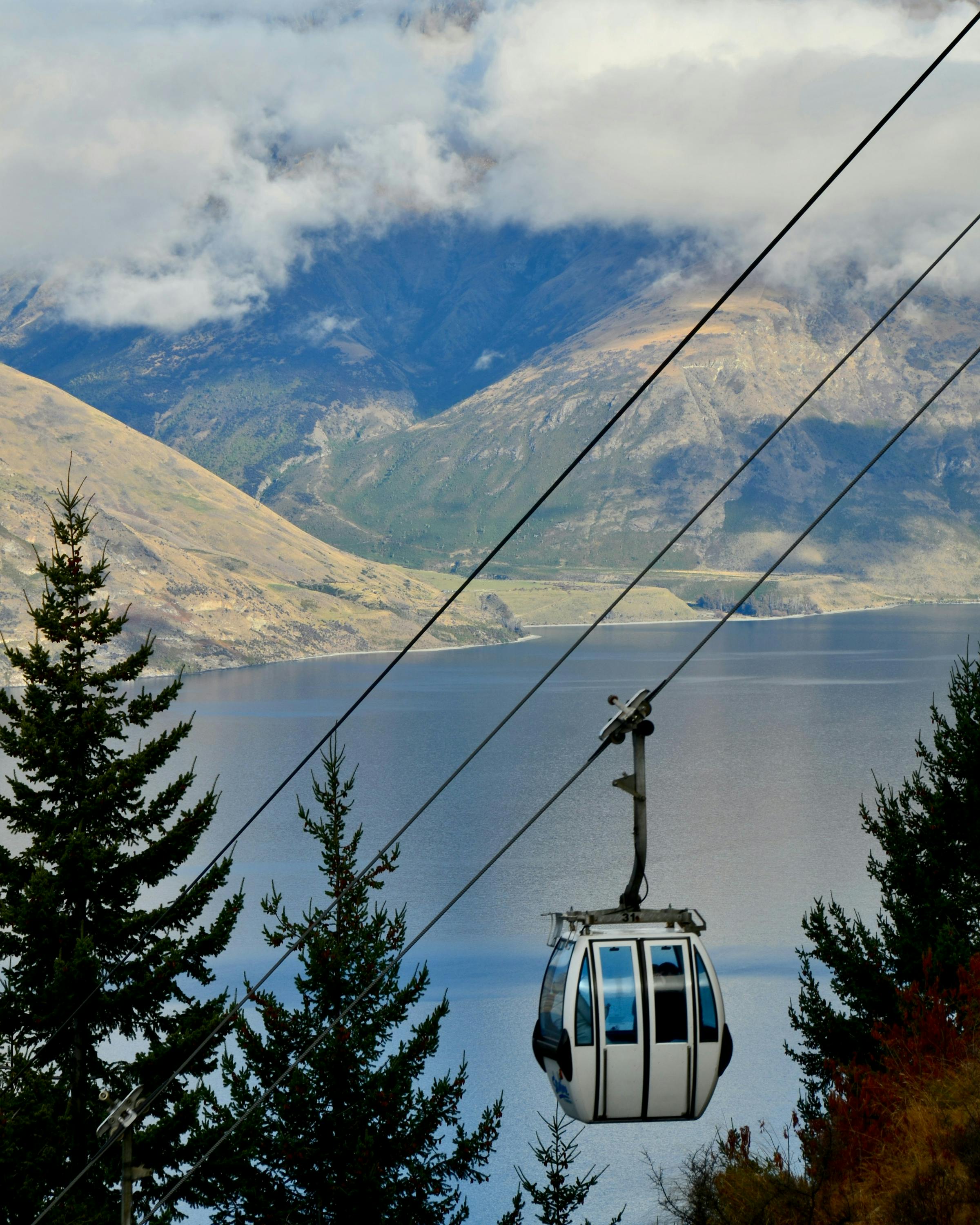 A gondola cabin rides above Queenstown with Lake Wakatipu and mountains behind, low clouds drifting across peaks.