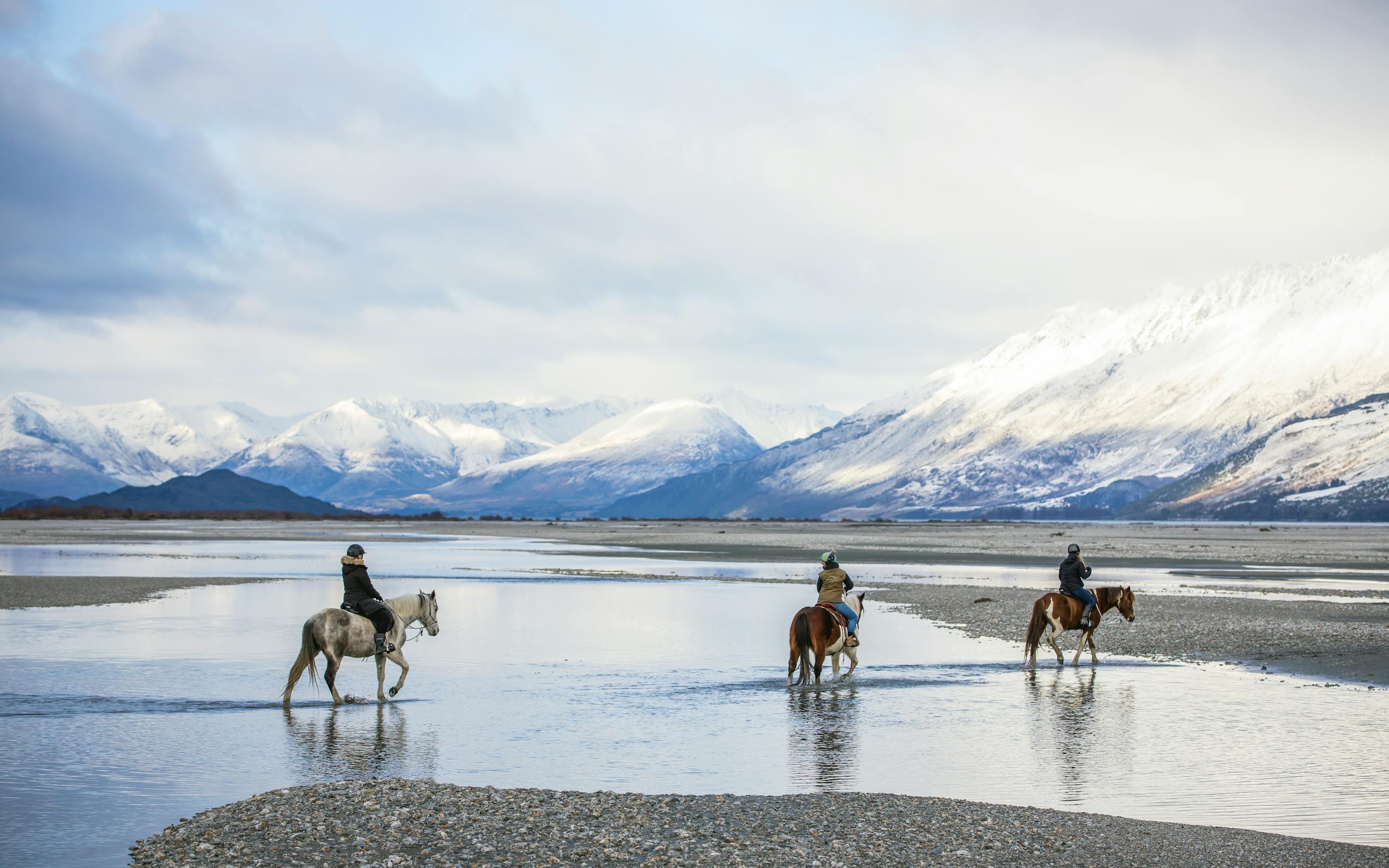 Three horseback riders cross a reflective shoreline near Glenorchy, with snowy peaks and soft light beyond.