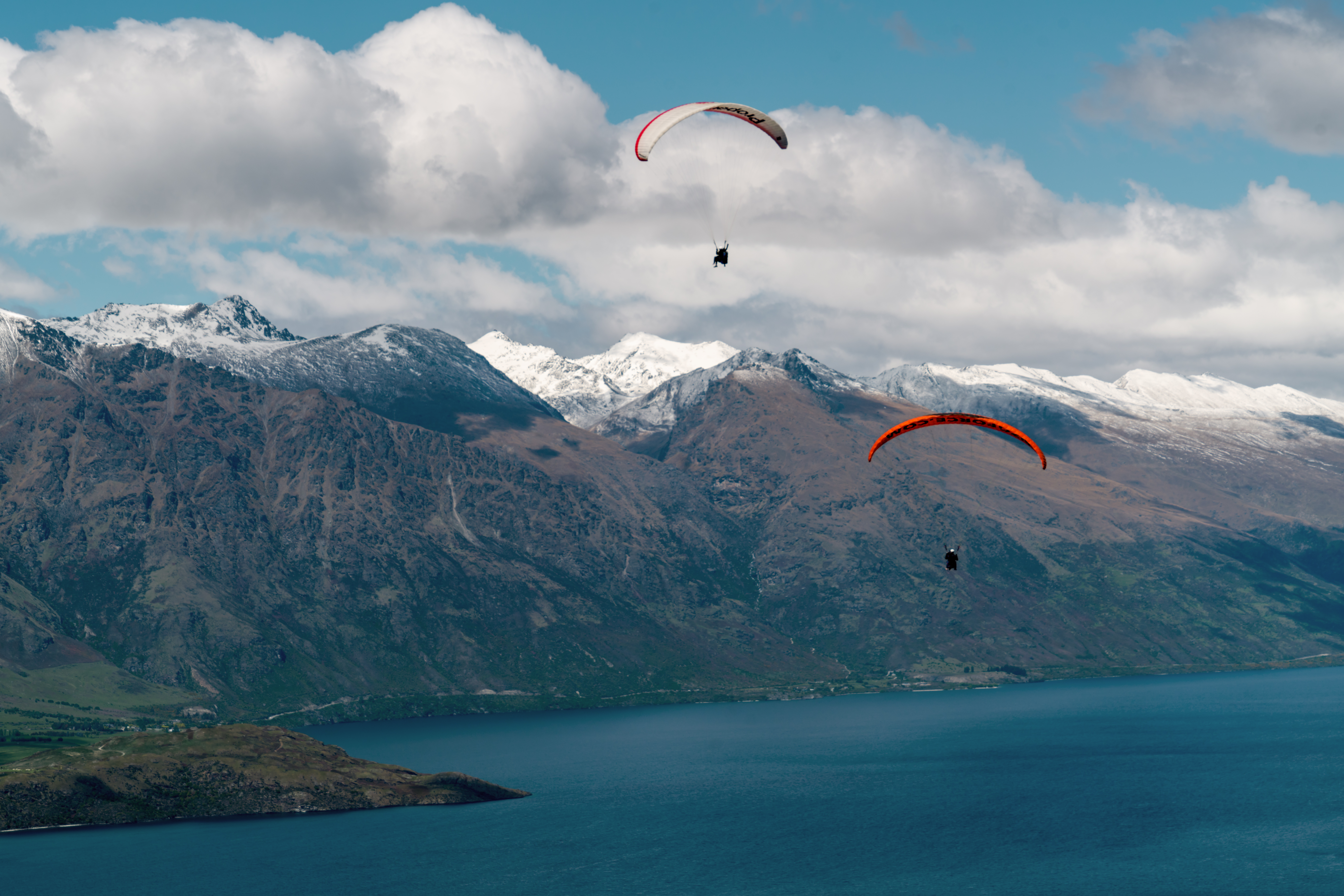 Two paragliders float above a blue lake and dark mountains, with snowy peaks rising beneath thick white clouds.