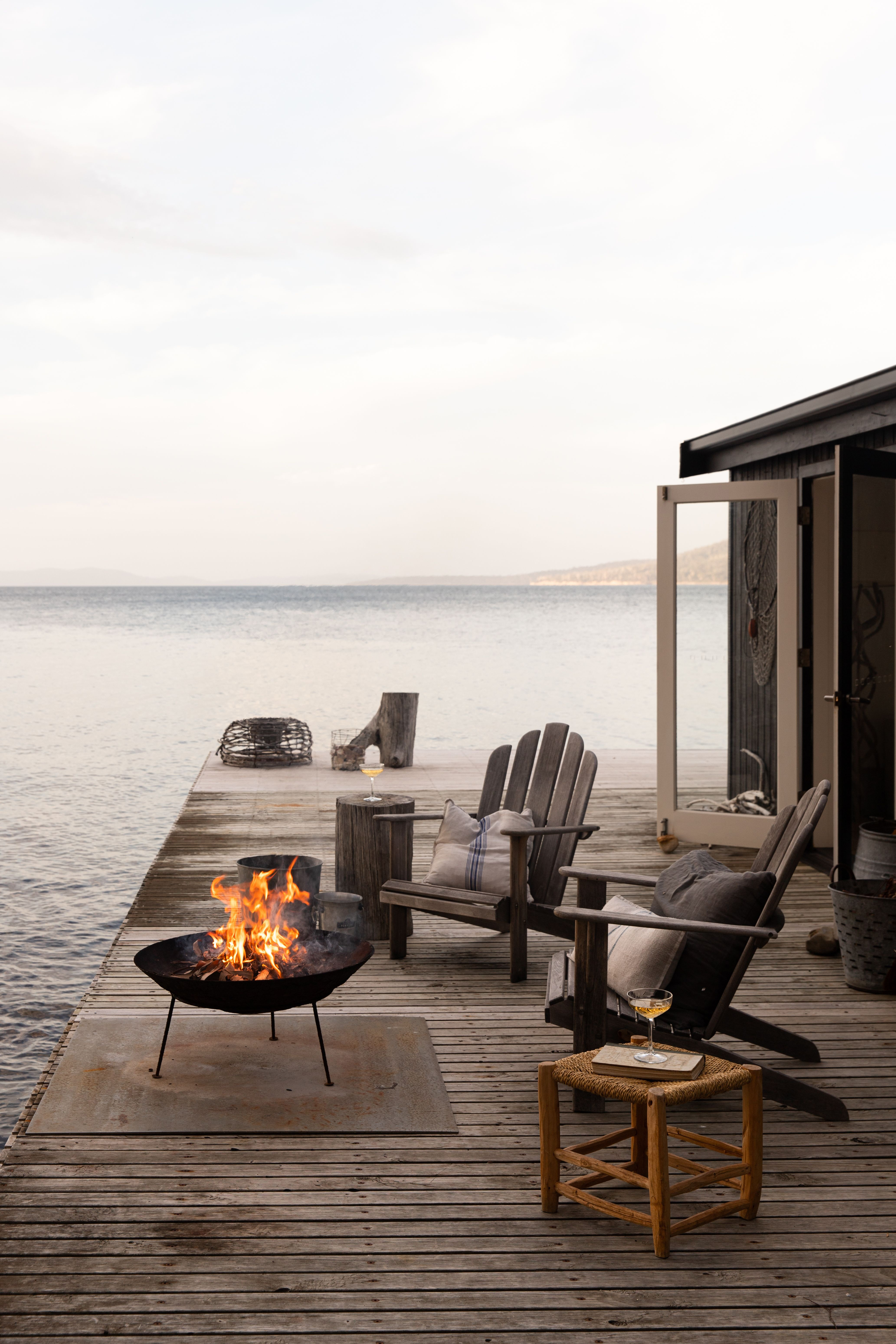 Outdoor chairs sit on a deck beside the ocean, with a fire pit glowing and waves lapping under a pale sky.
