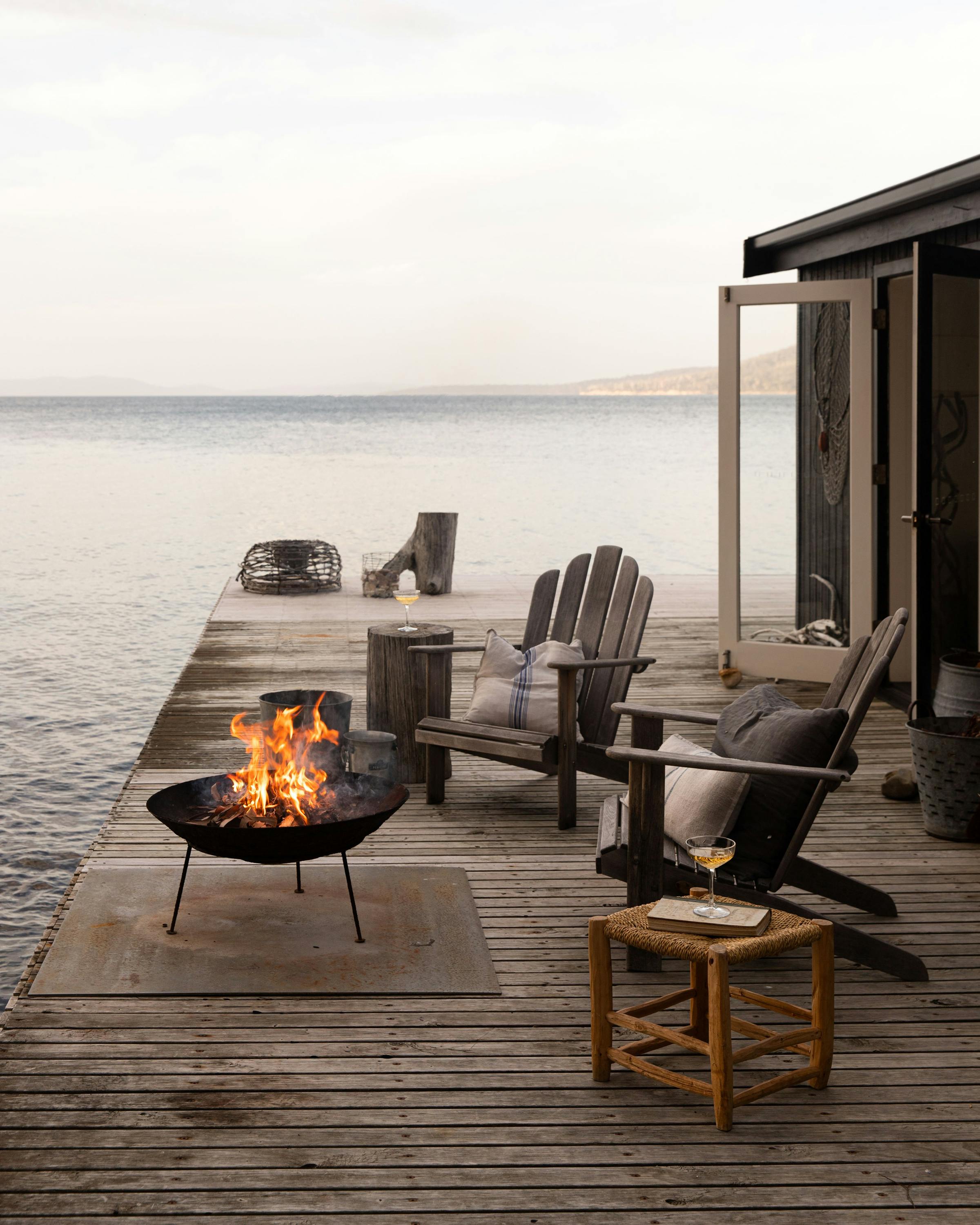 Outdoor chairs sit on a deck beside the ocean, with a fire pit glowing and waves lapping under a pale sky.