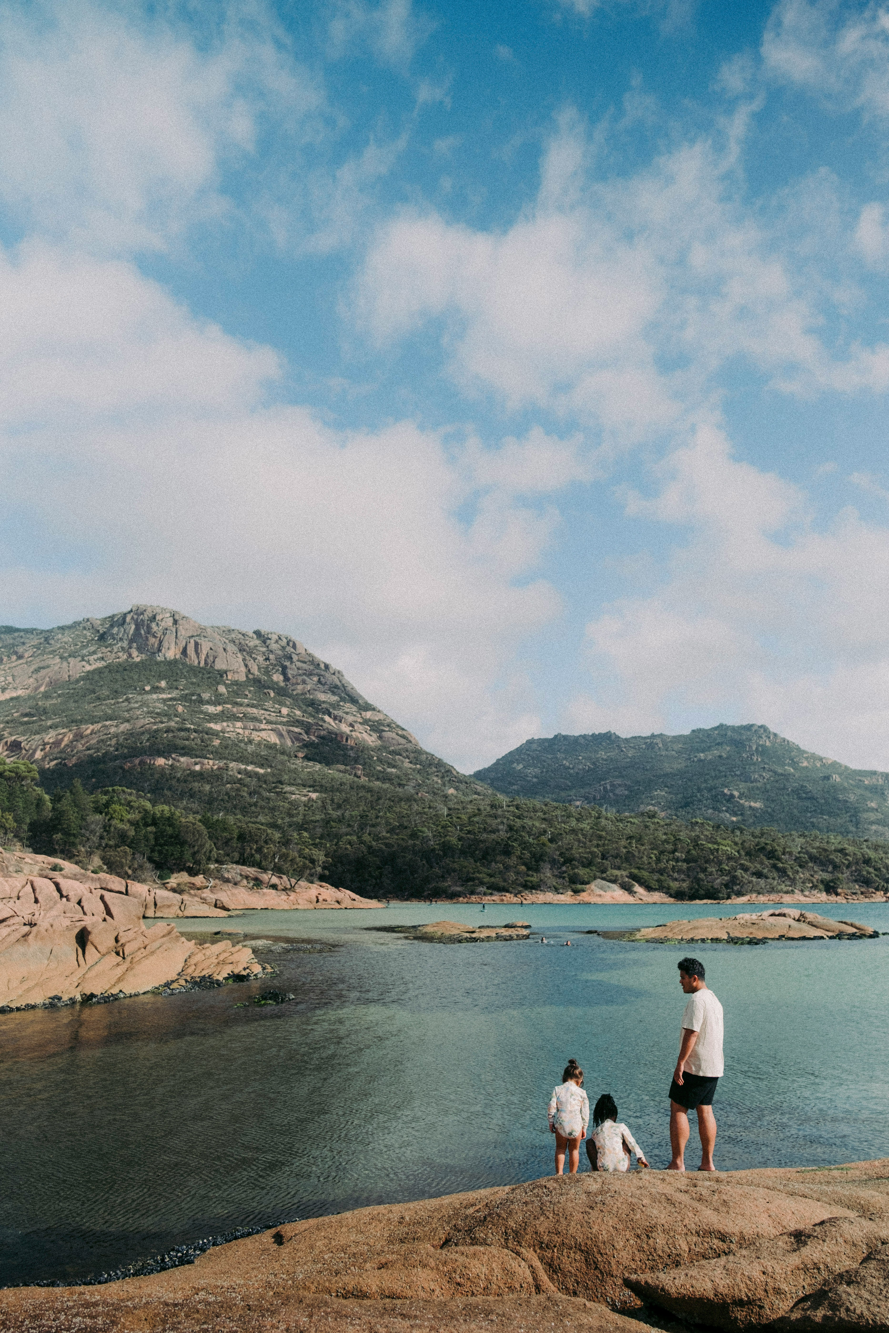 Three people stand on rocky shore by a calm inlet, looking toward low mountains beneath blue sky and clouds.