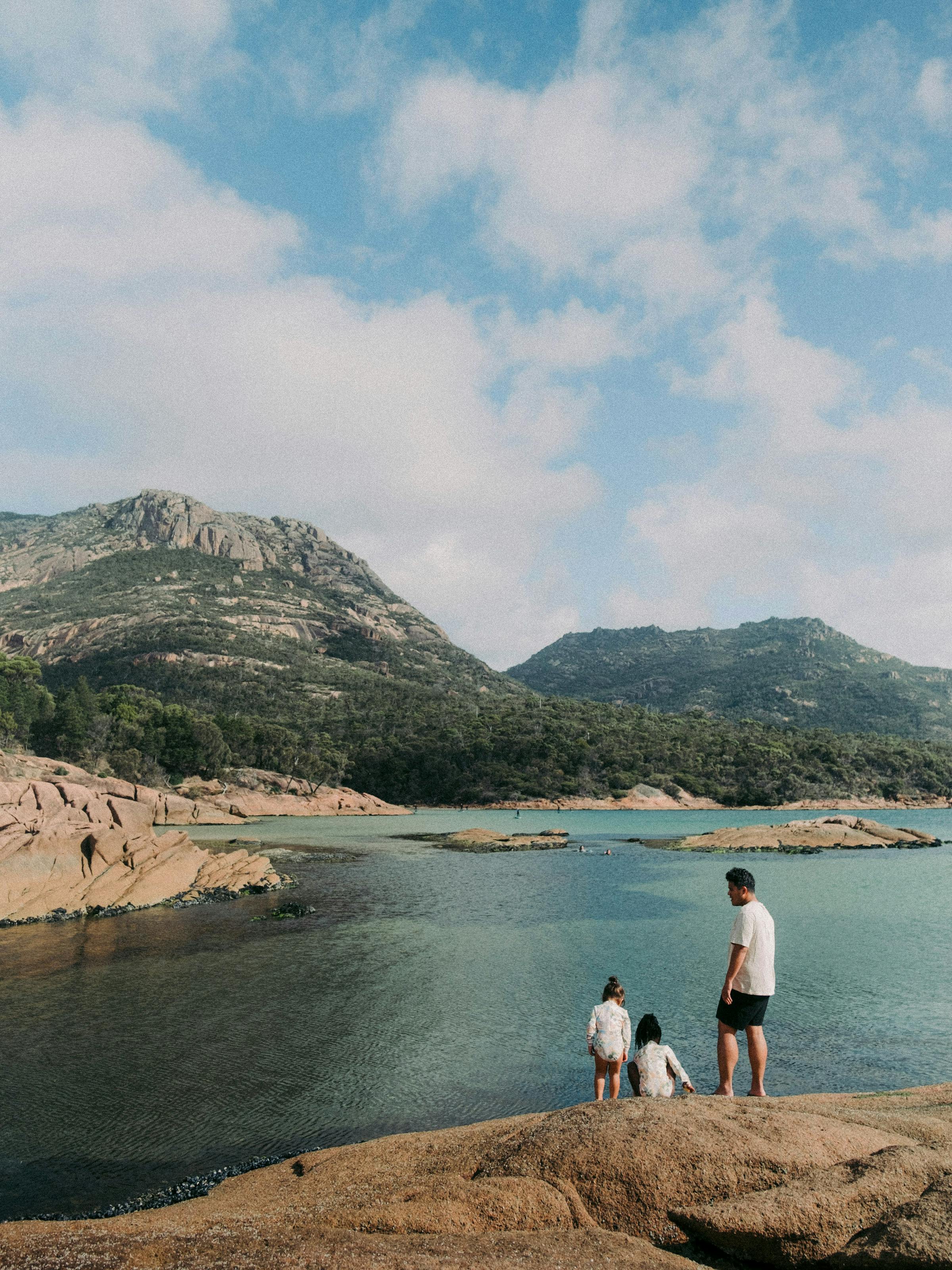 Three people stand on rocky shore by a calm inlet, looking toward low mountains beneath blue sky and clouds.