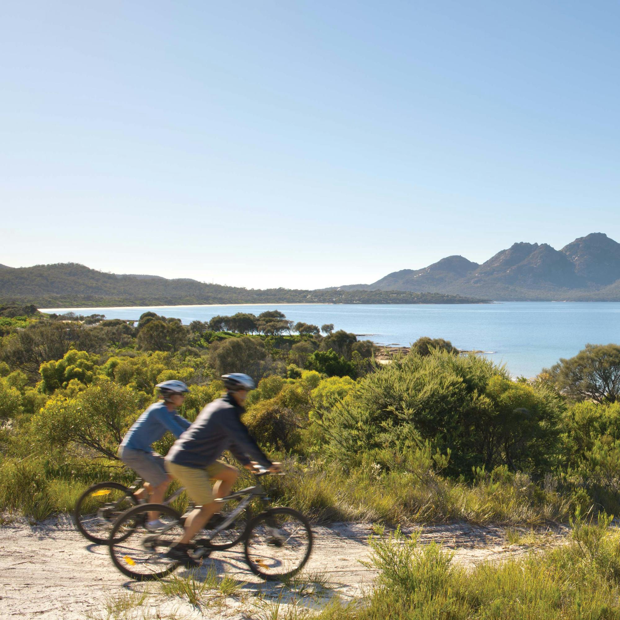 Two mountain bikers ride along a sandy trail near Freycinet, with blue bays and rugged peaks in the distance.