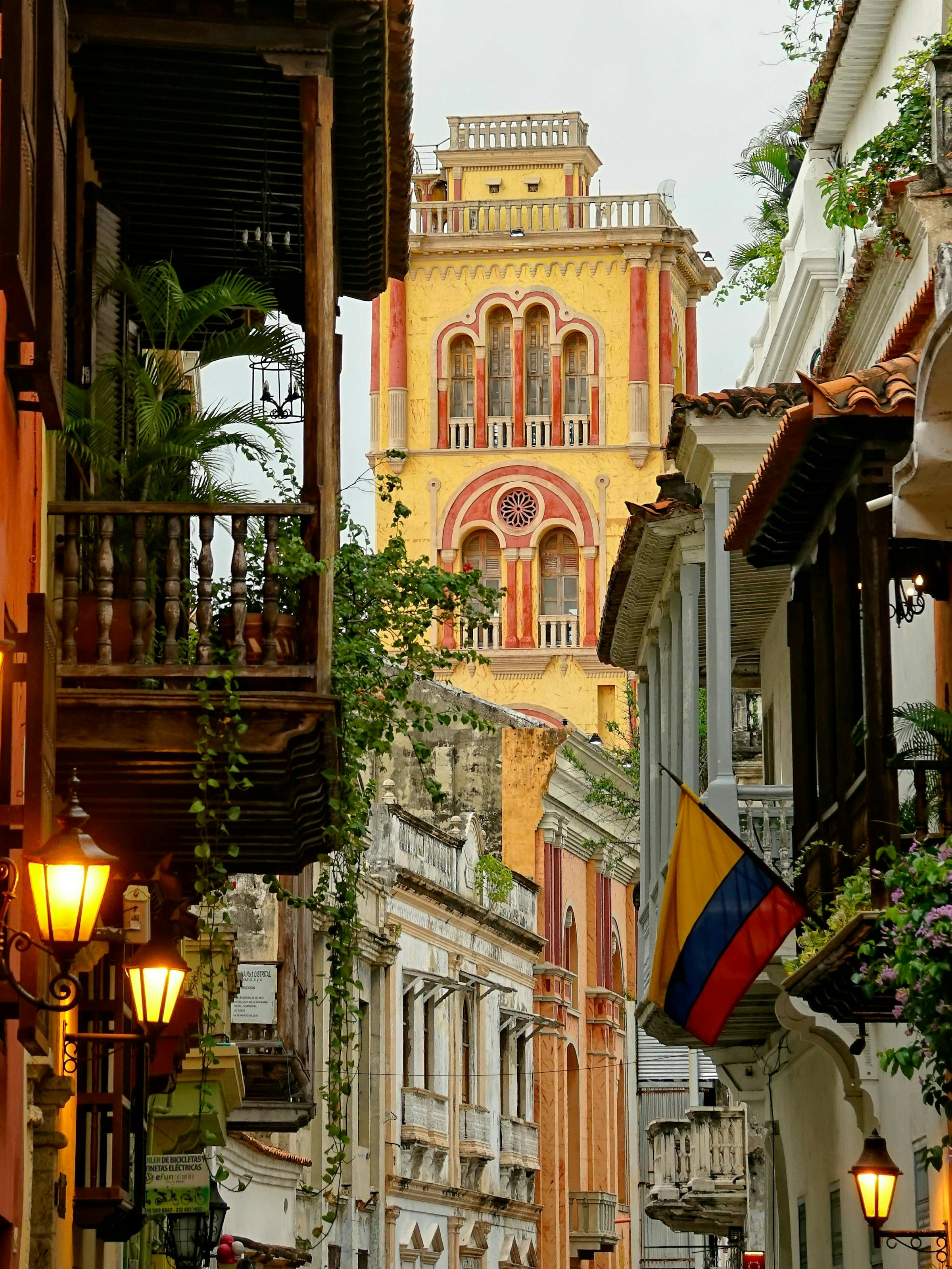 A narrow Cartagena street is lined with balconies and lanterns, with the Colombian flag hanging near a church tower.