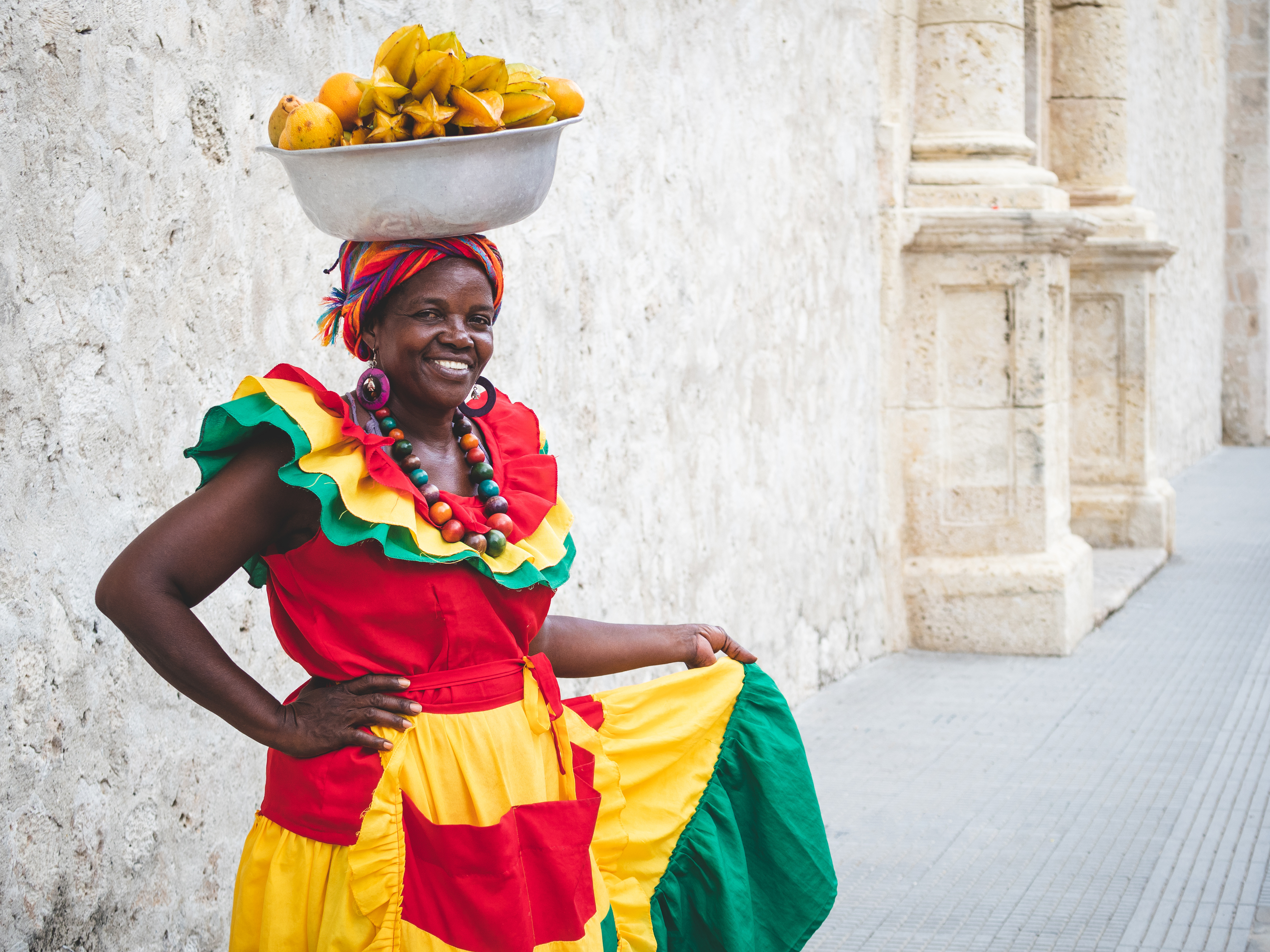 A smiling palenquera in bright dress balances a bowl of fruit on her head beside a stone wall in Cartagena.