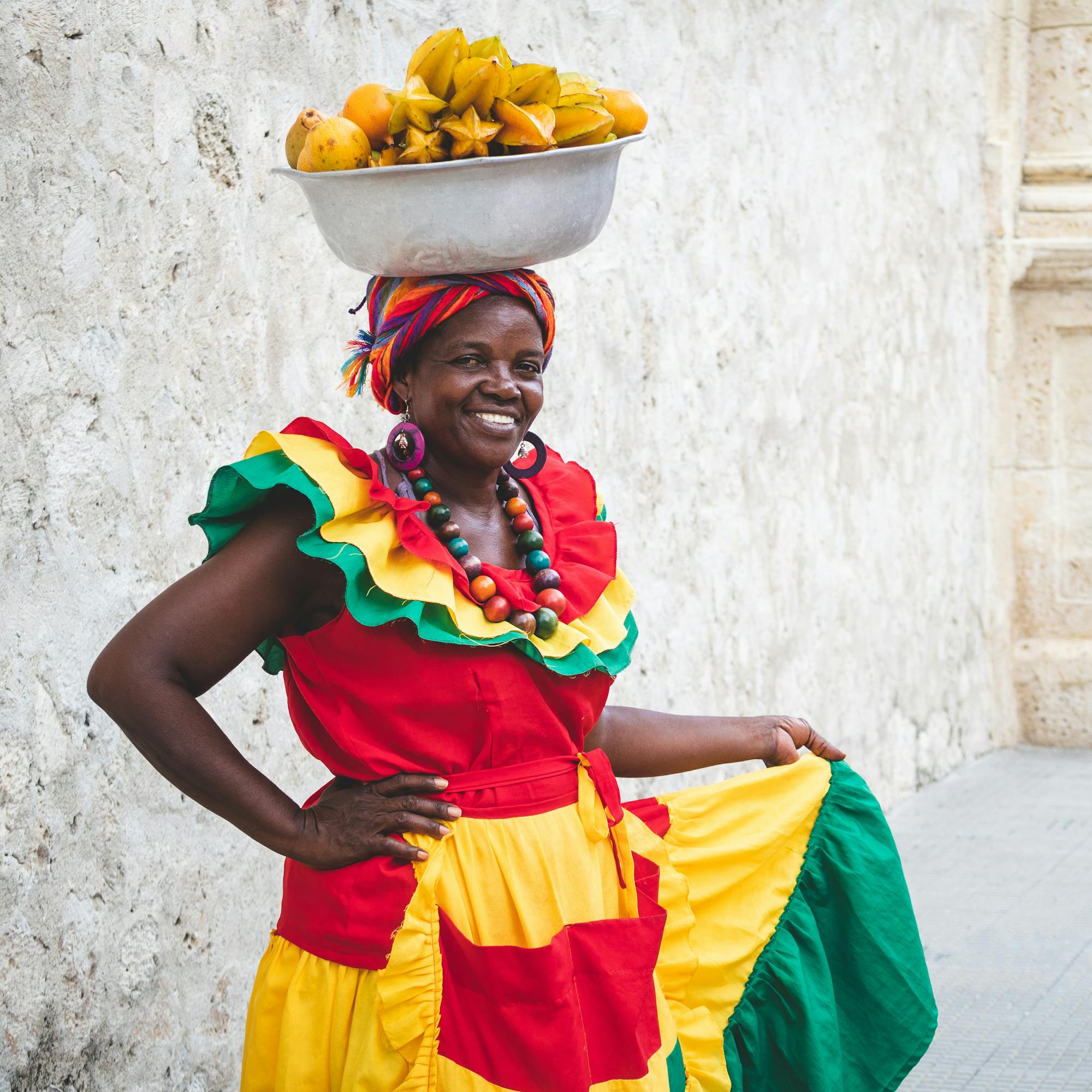 A smiling palenquera in bright dress balances a bowl of fruit on her head beside a stone wall in Cartagena.