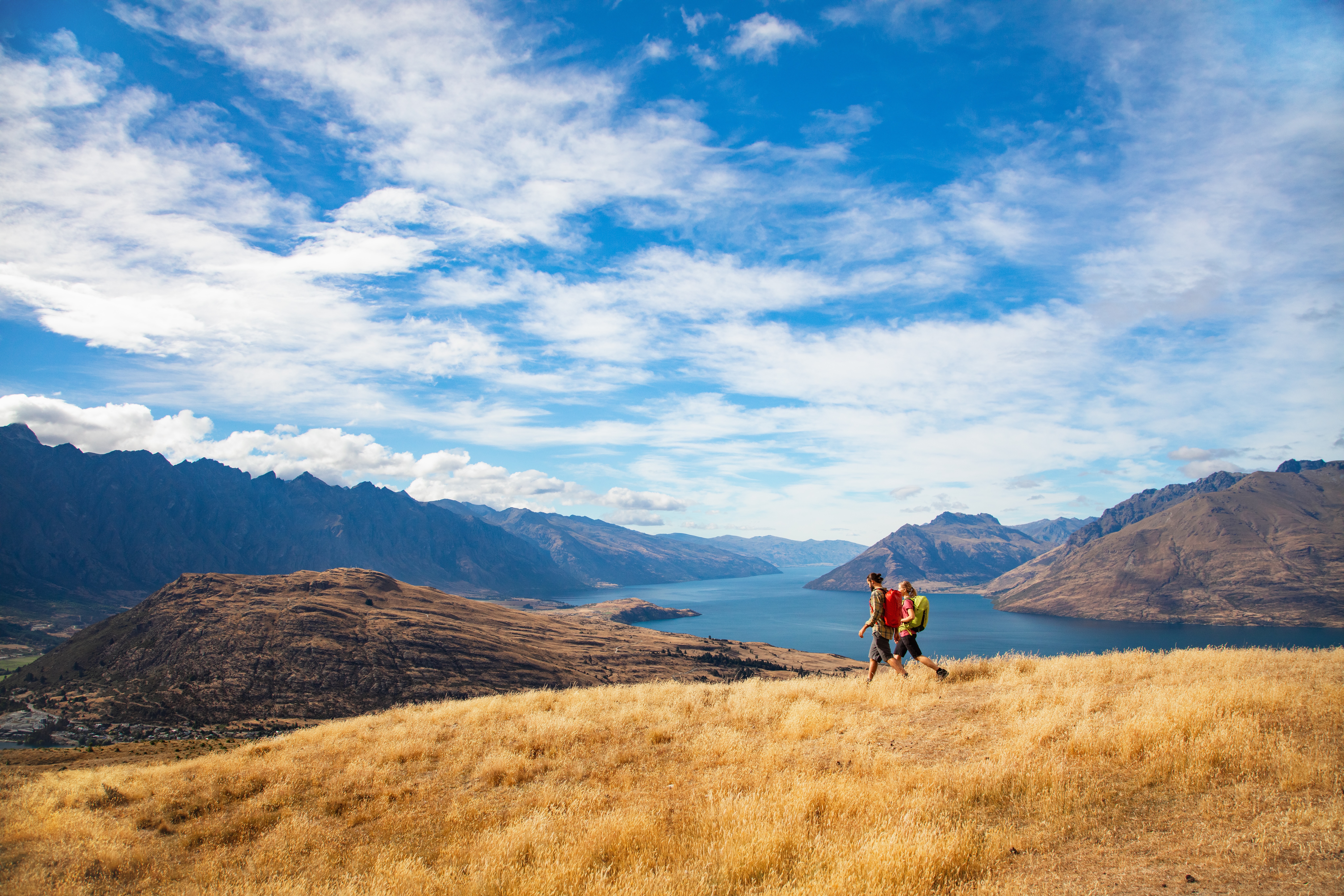 Two hikers stand on golden tussock above Lake Wakatipu, with rugged peaks and dramatic clouds overhead.