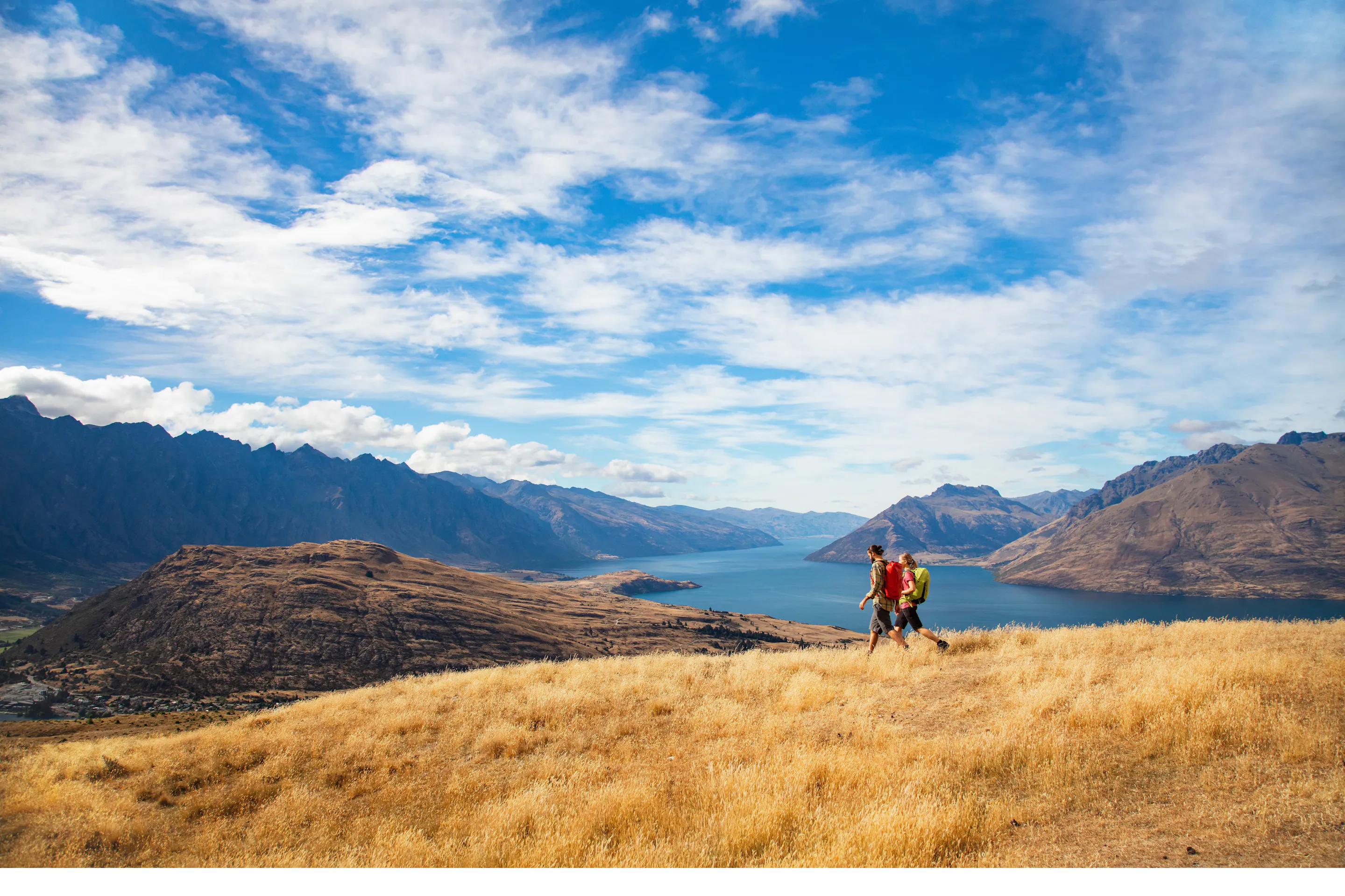 Two hikers stand on golden tussock above Lake Wakatipu, with rugged peaks and dramatic clouds overhead.