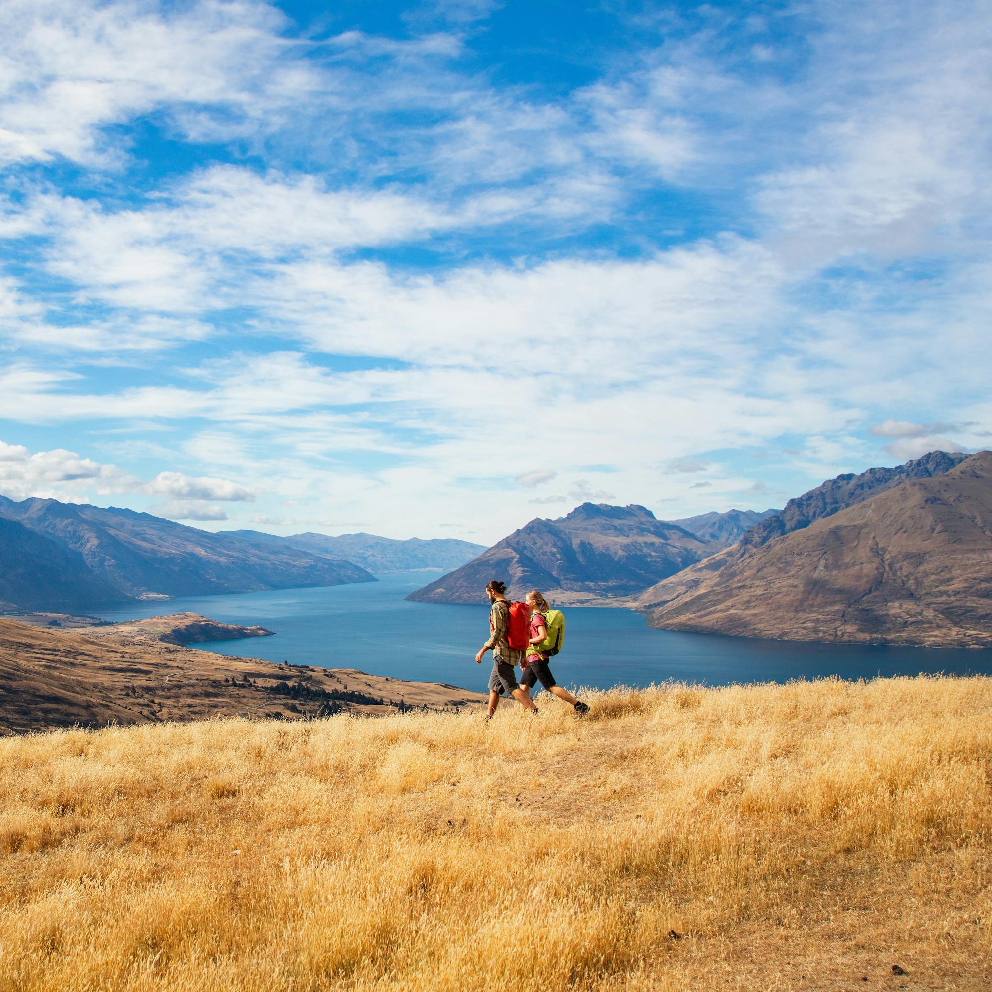 Two hikers stand on golden tussock above Lake Wakatipu, with rugged peaks and dramatic clouds overhead.