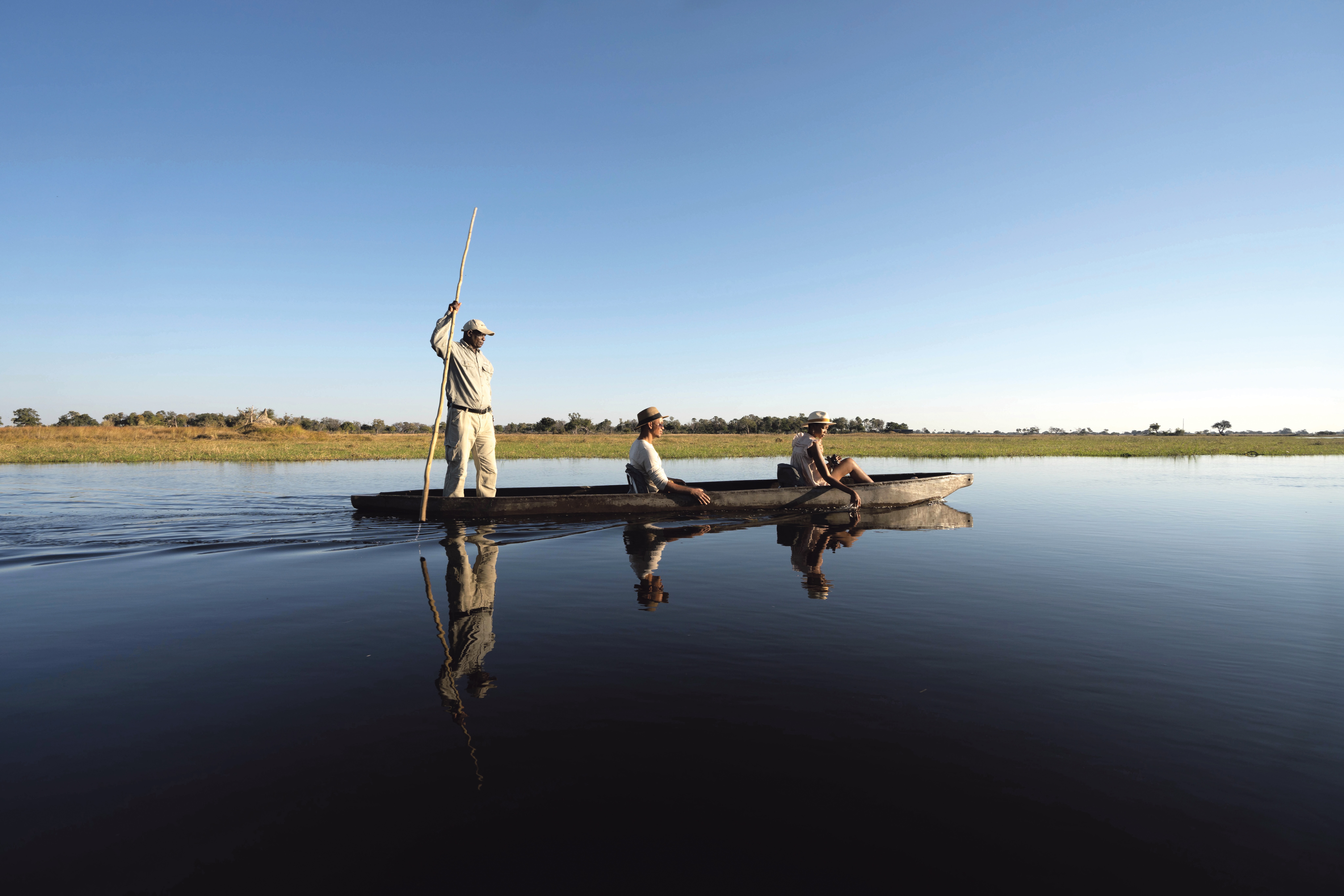 A mokoro poler stands at the stern as the canoe drifts across still water, mirrored beneath a wide blue sky.