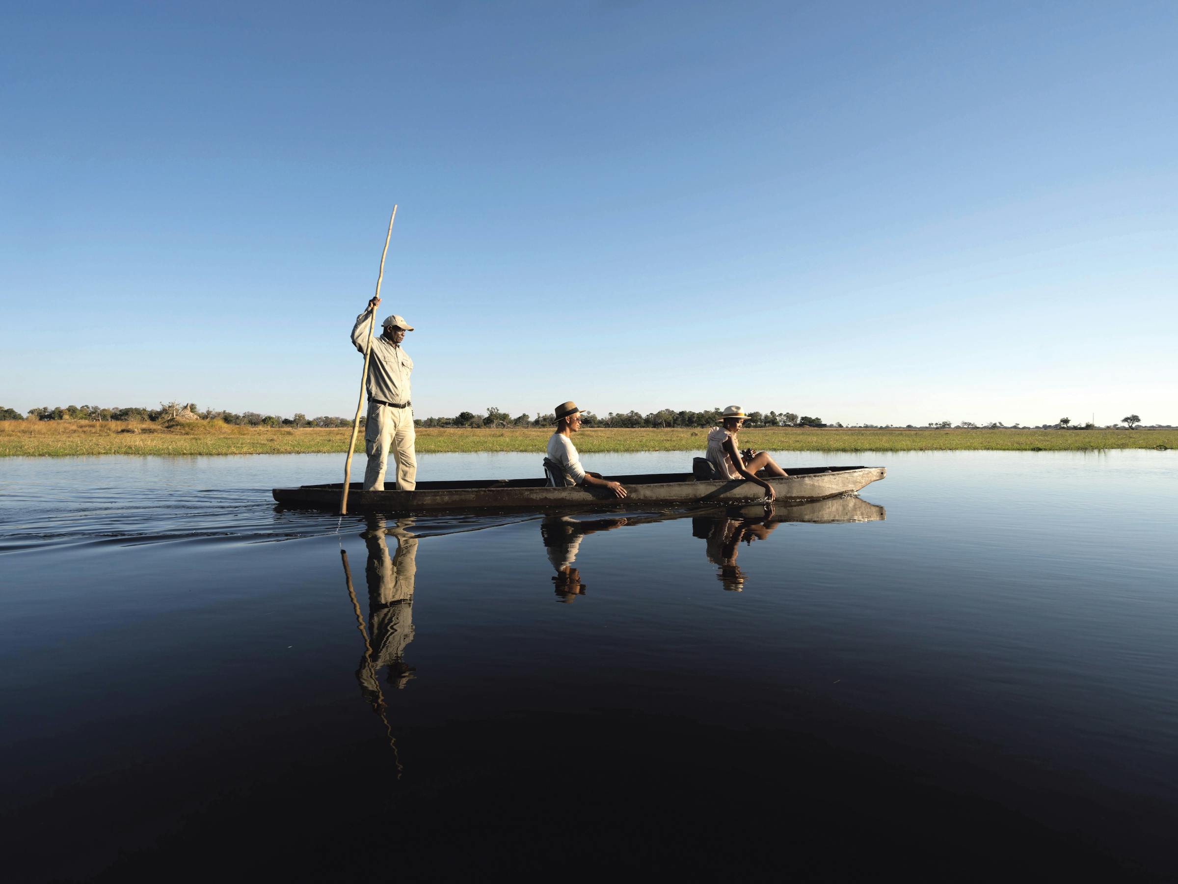 A mokoro poler stands at the stern as the canoe drifts across still water, mirrored beneath a wide blue sky.