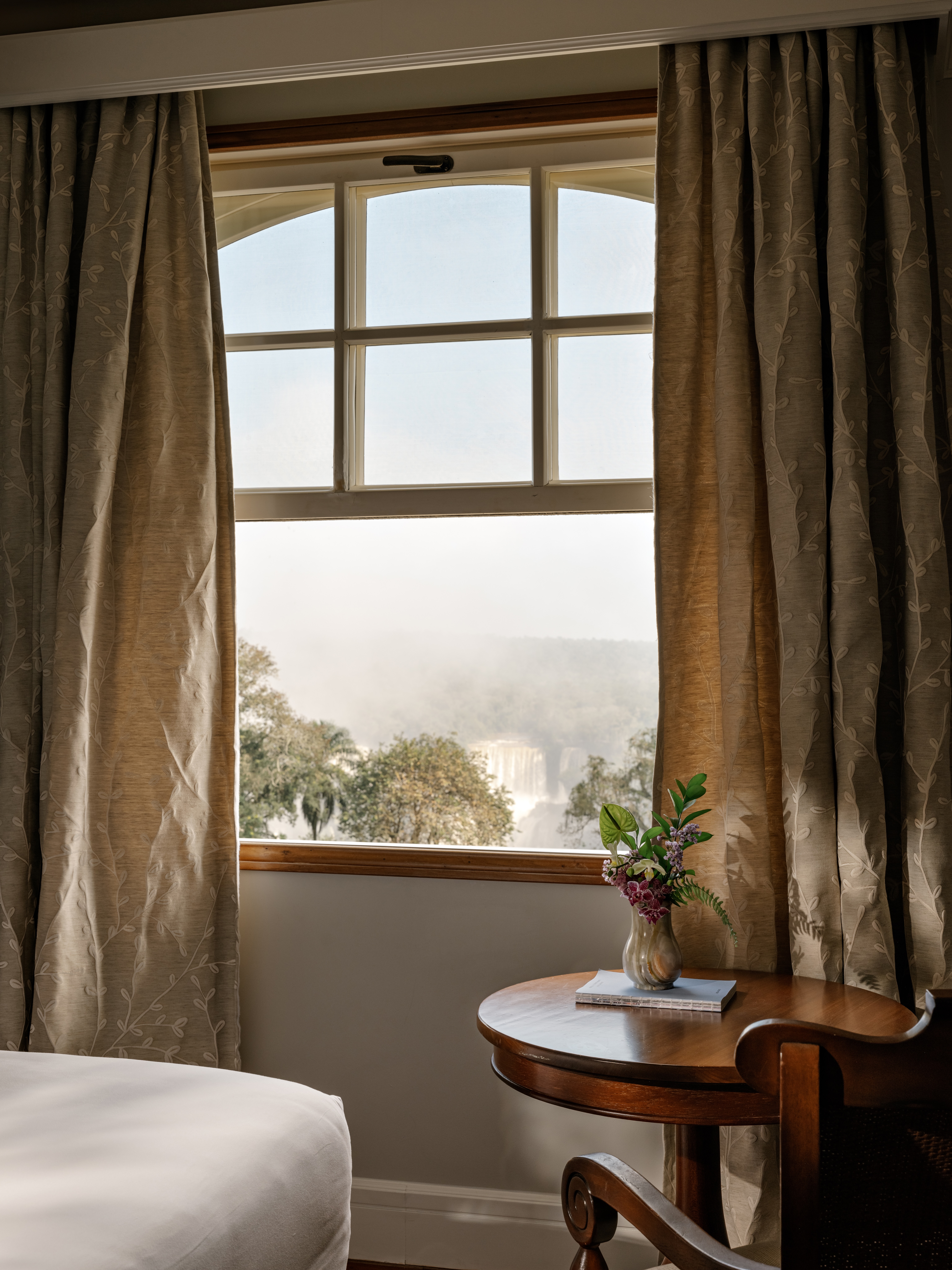 Inside a guest room, sunlight falls through a window onto a chair and table, with trees visible beyond curtains.