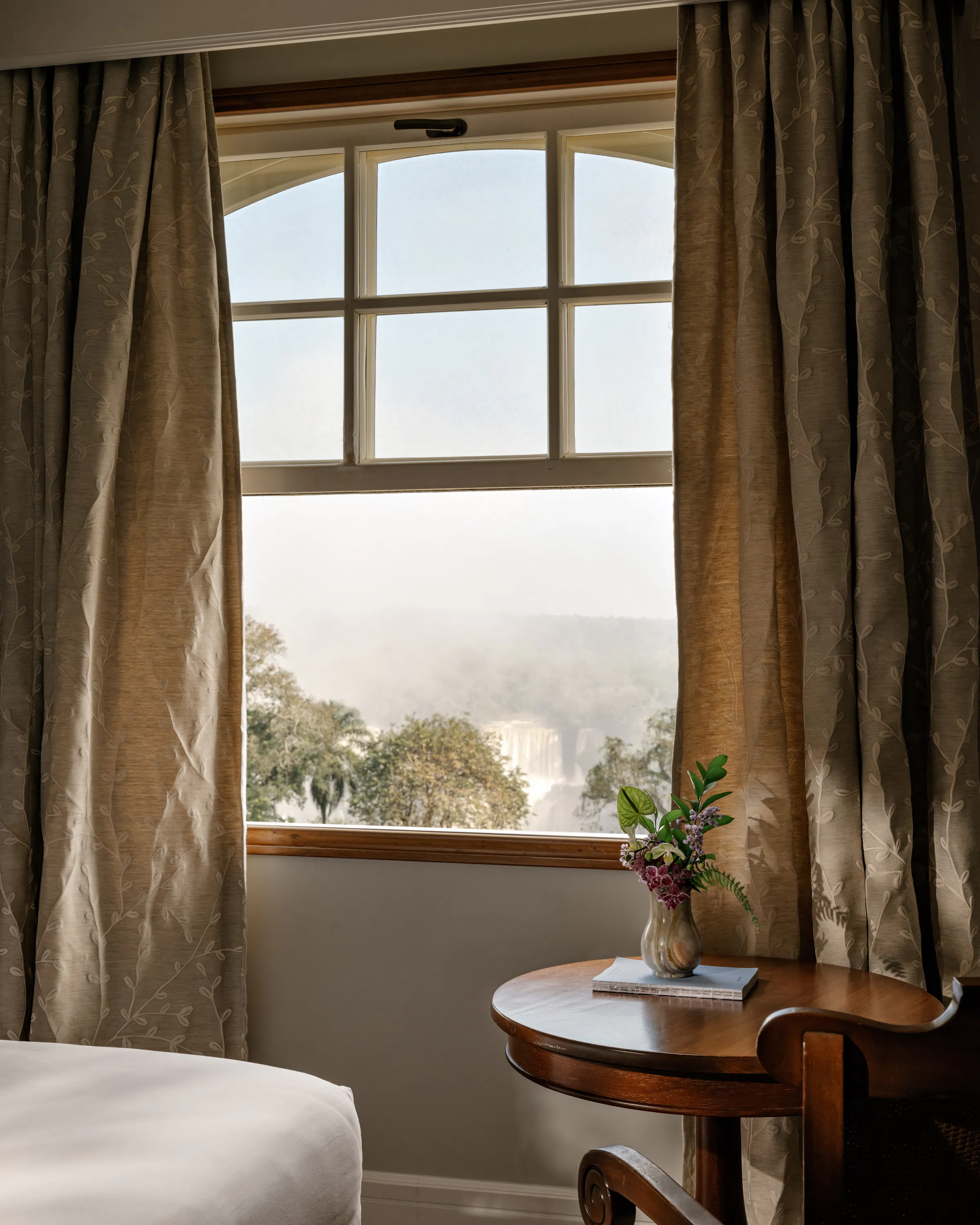 Inside a guest room, sunlight falls through a window onto a chair and table, with trees visible beyond curtains.