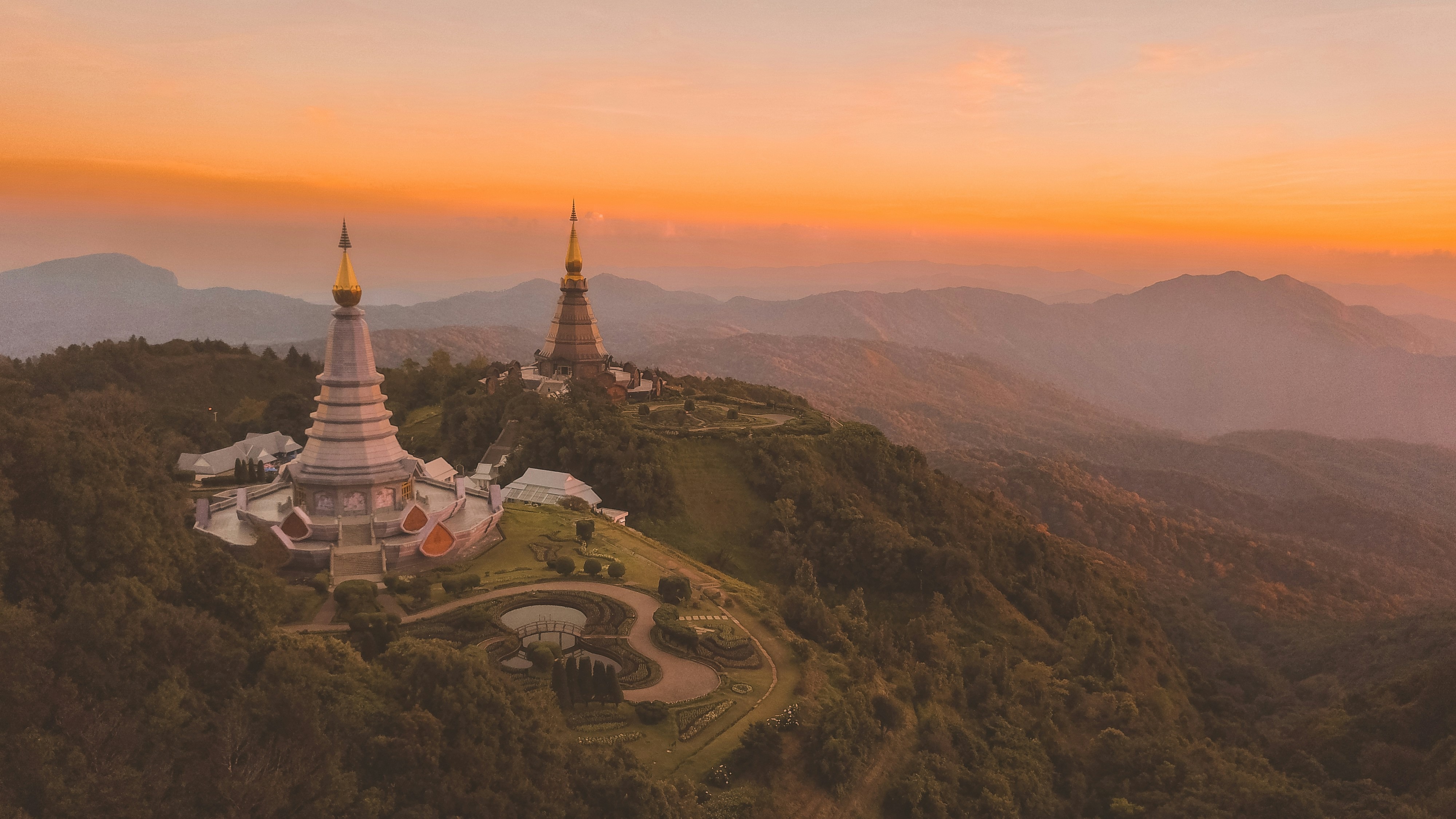 Two ornate temple pagodas with golden spires rise above a lush, mountainous landscape at sunset. The sky glows in shades of orange and pink, casting a peaceful light over the hills and surrounding forest.