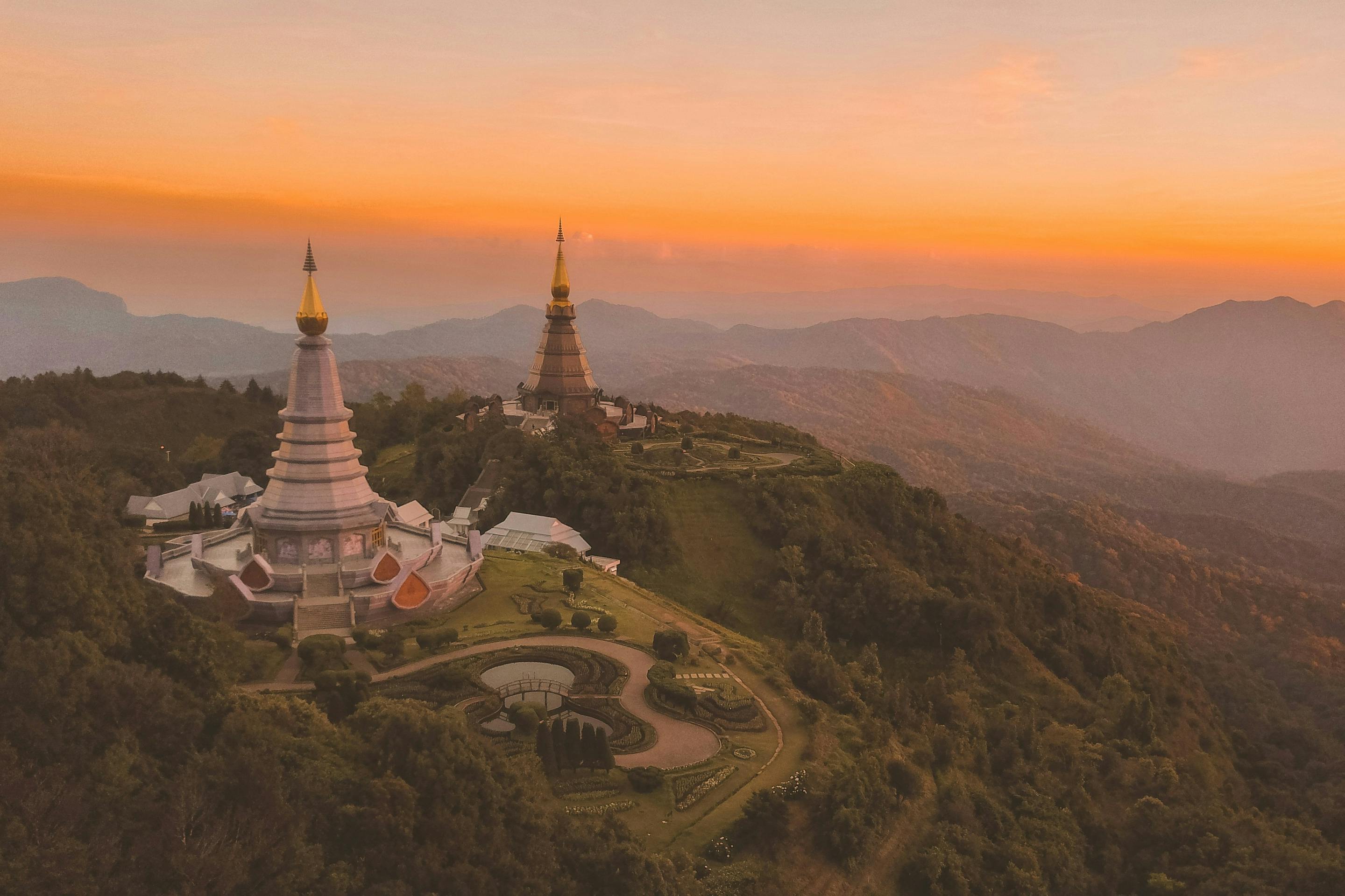 Two ornate temple pagodas with golden spires rise above a lush, mountainous landscape at sunset. The sky glows in shades of orange and pink, casting a peaceful light over the hills and surrounding forest.