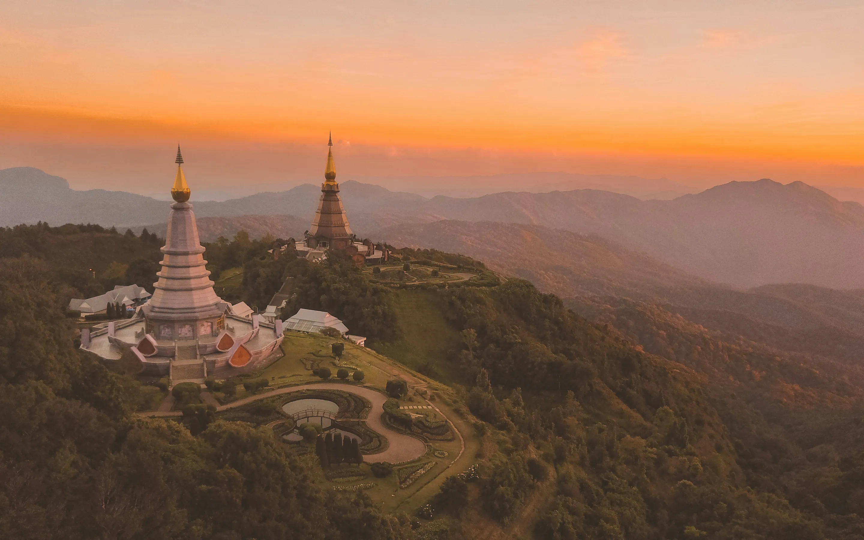 Two ornate temple pagodas with golden spires rise above a lush, mountainous landscape at sunset. The sky glows in shades of orange and pink, casting a peaceful light over the hills and surrounding forest.