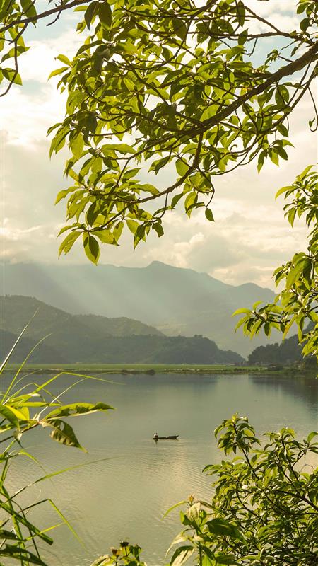 A calm lake is framed by leafy green branches in the foreground. A small boat floats on the water, with misty mountains layered in the distance under a soft, hazy sky.