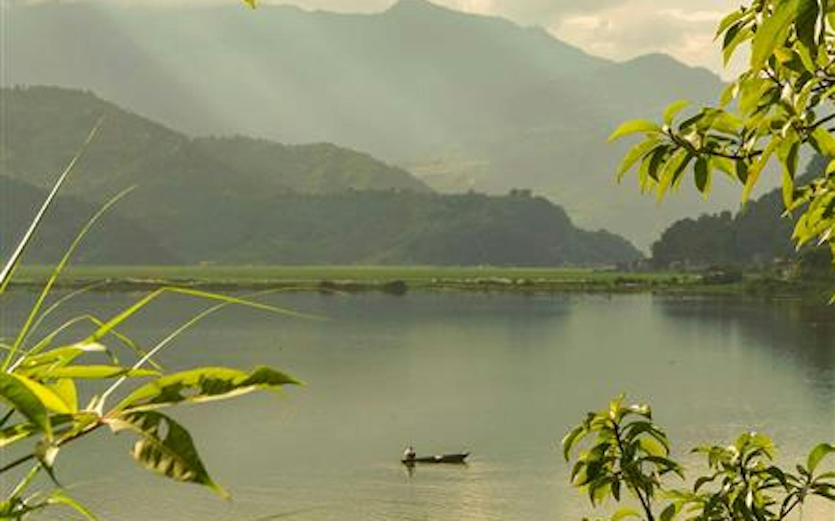 A calm lake is framed by leafy green branches in the foreground. A small boat floats on the water, with misty mountains layered in the distance under a soft, hazy sky.