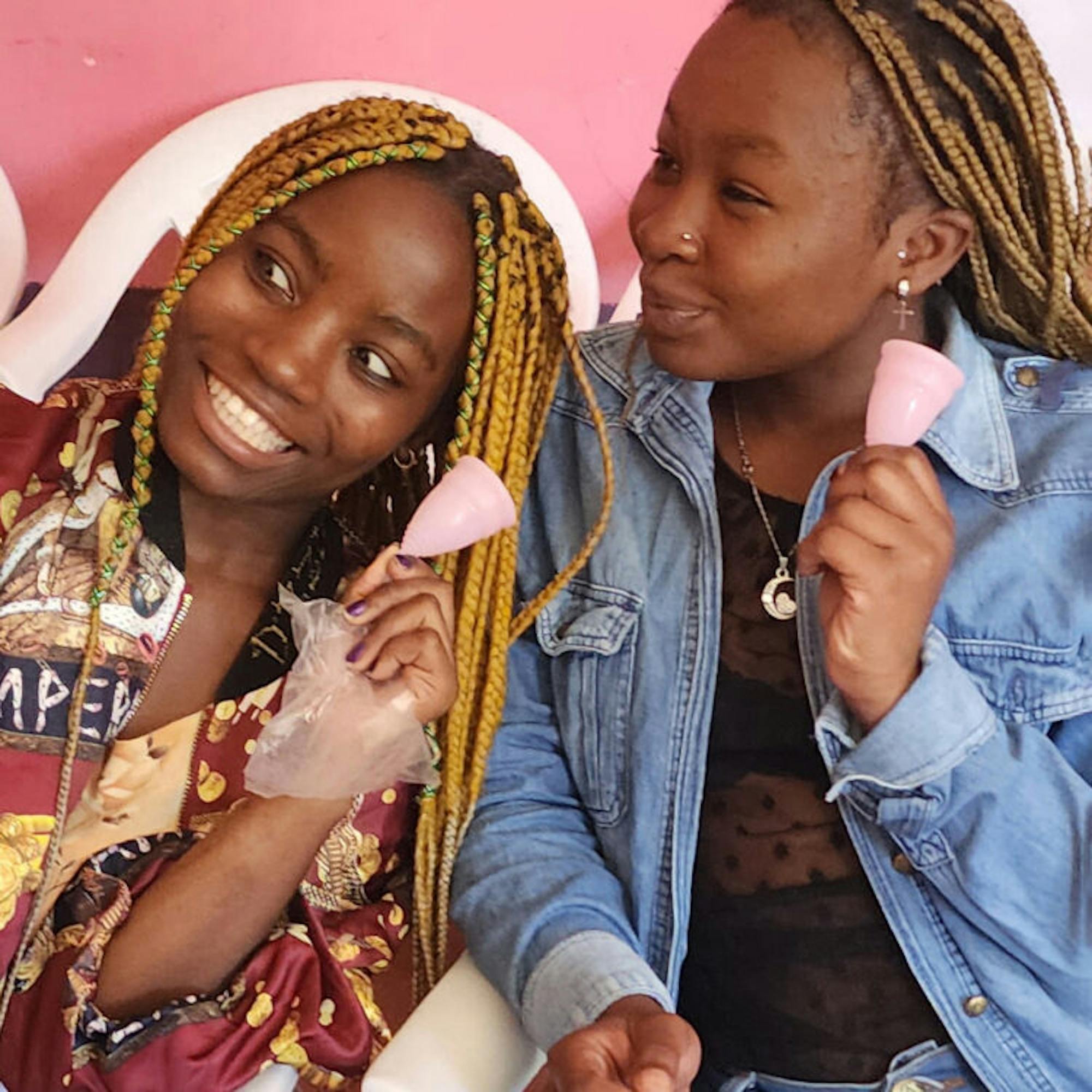 Two women smiling together hold pink menstrual cups against a pink background.