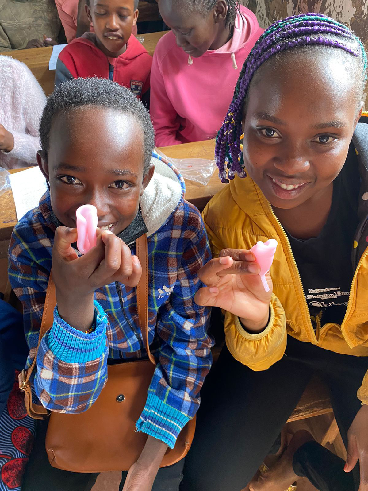Students in a classroom hold pink hygiene items and worksheets while smiling toward the camera.