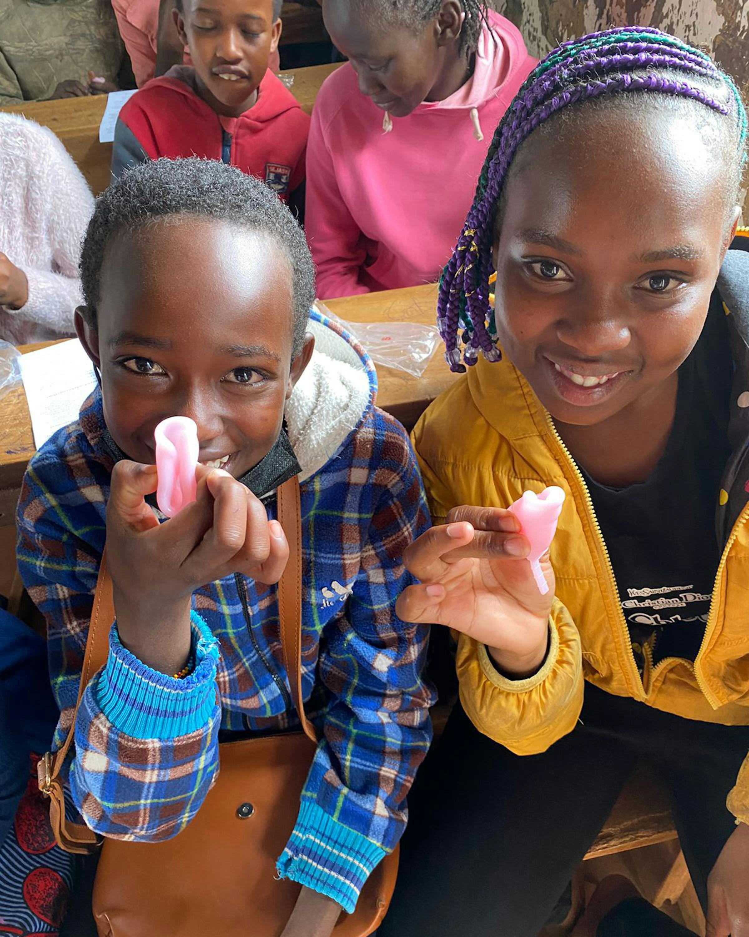 Students in a classroom hold pink hygiene items and worksheets while smiling toward the camera.
