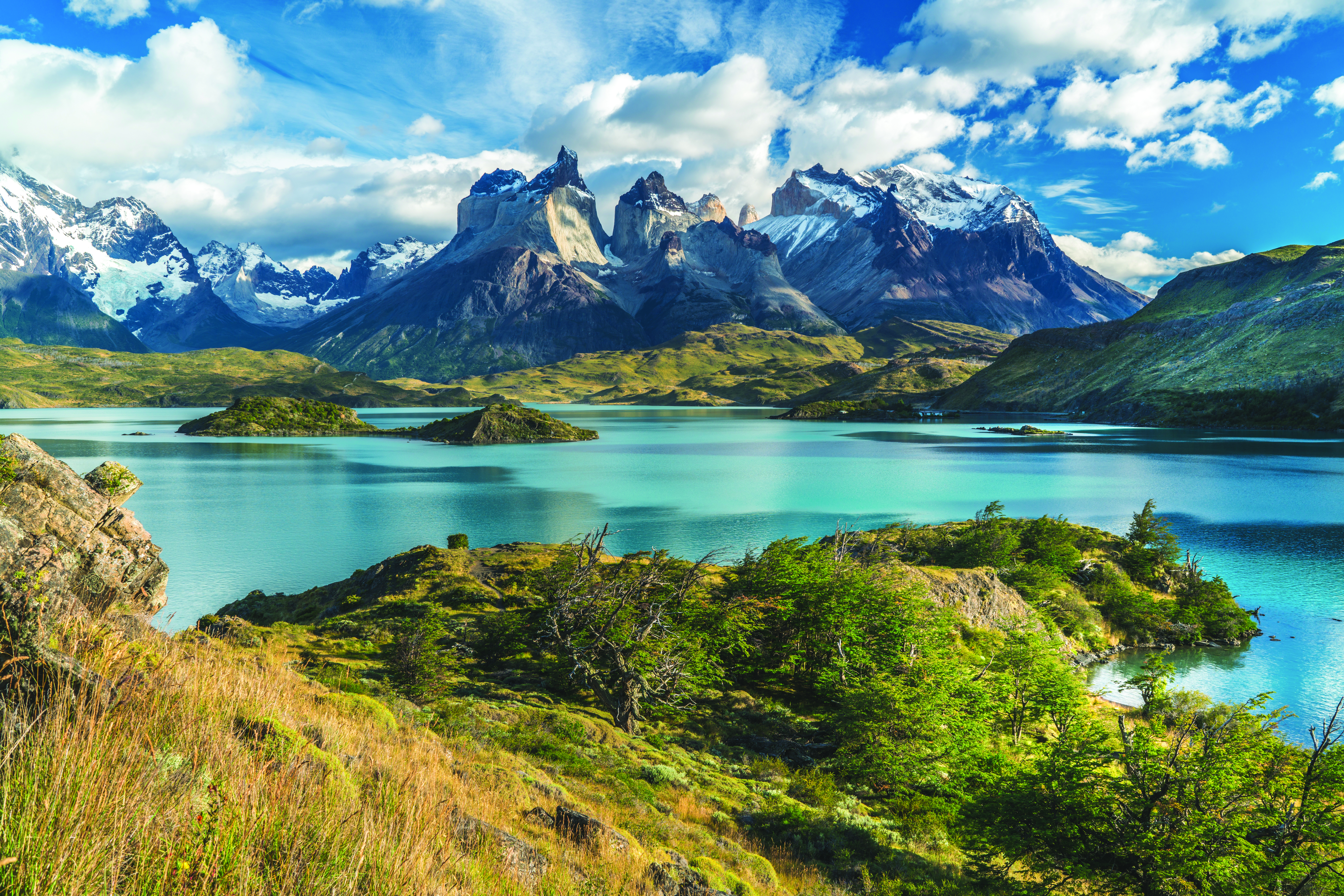 Jagged peaks rise above a calm lake and rocky shore in Torres del Paine, with wind-swept clouds overhead.