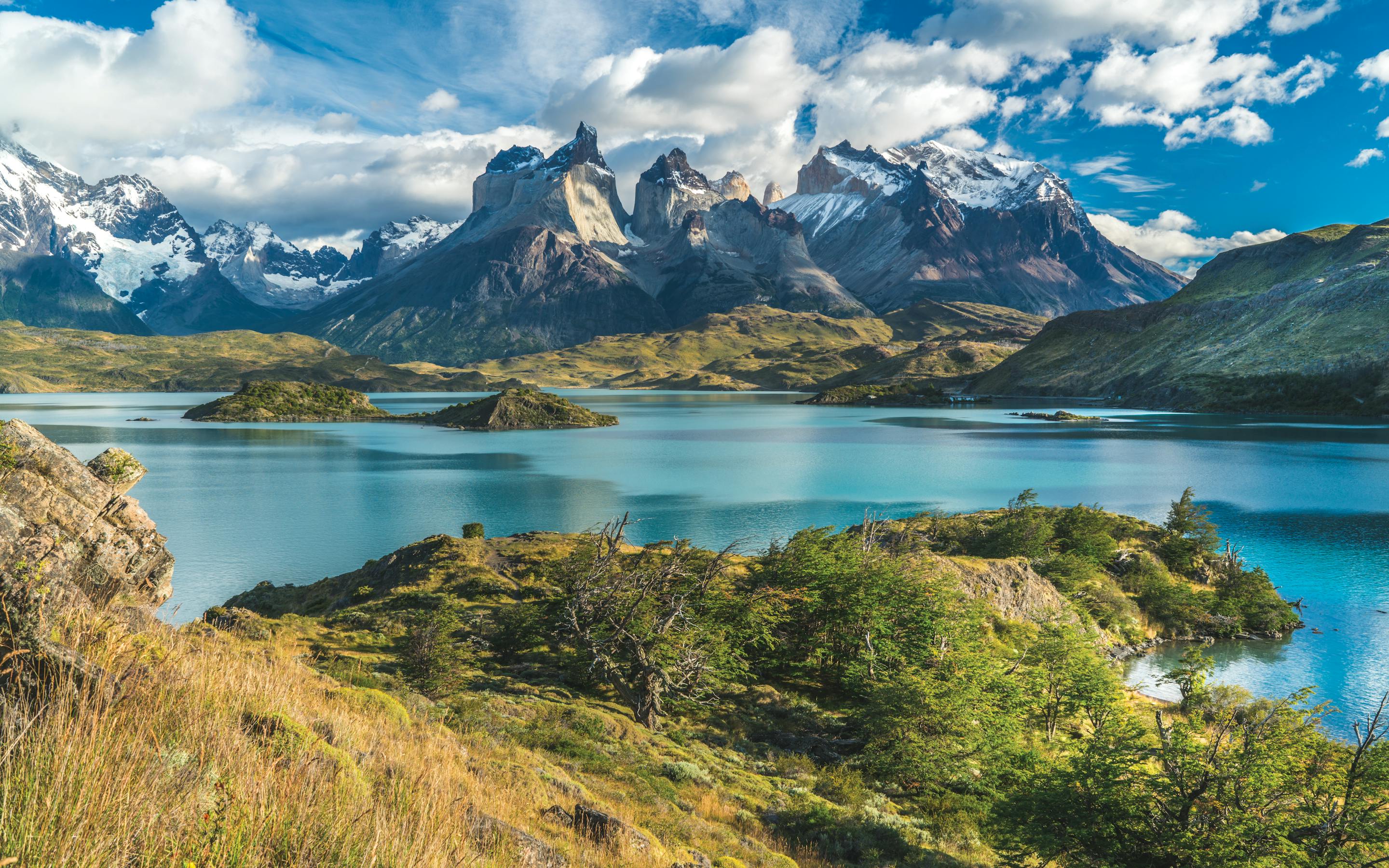 Jagged peaks rise above a calm lake and rocky shore in Torres del Paine, with wind-swept clouds overhead.