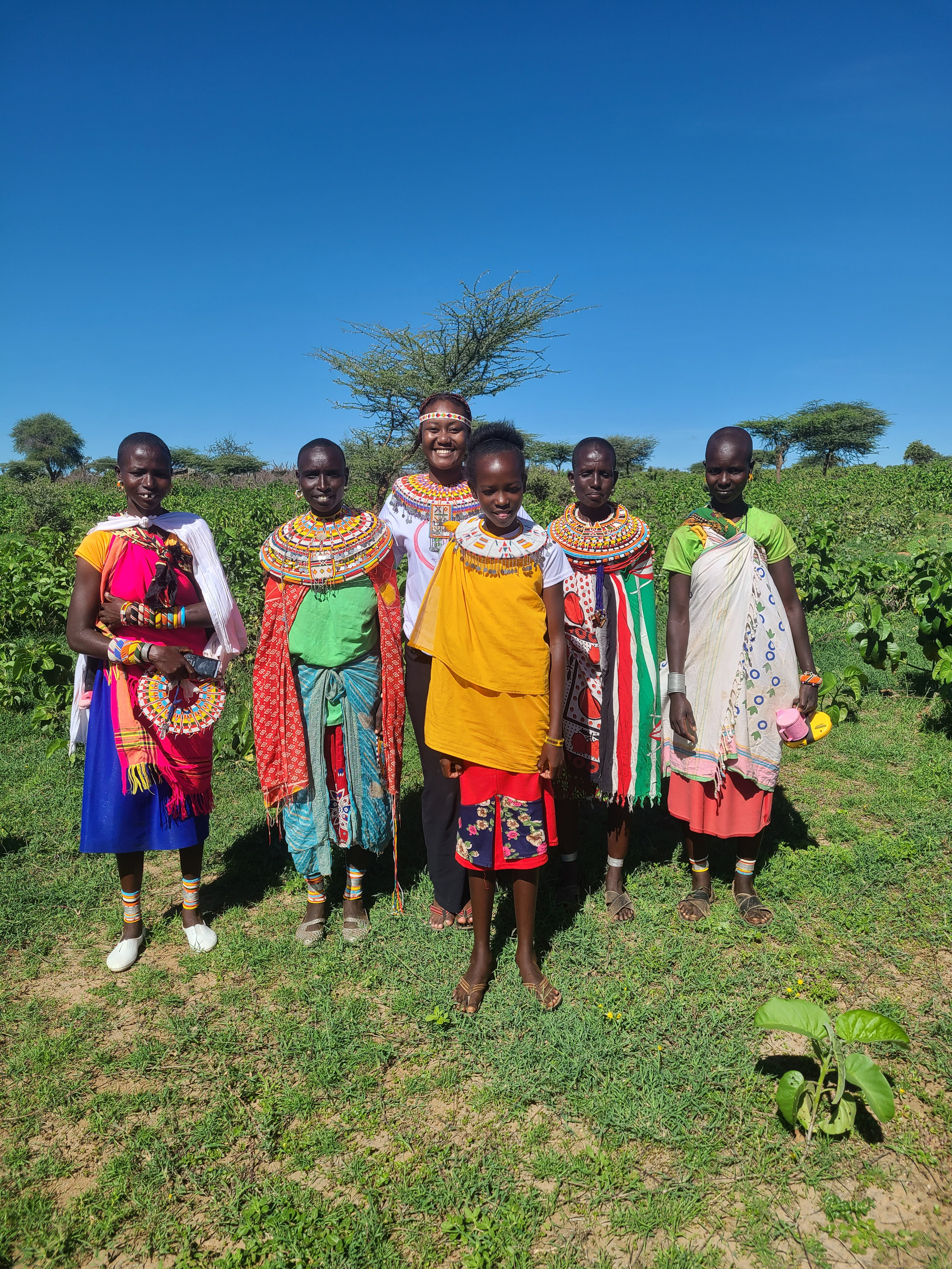 Five Samburu girls and women in beaded collars pose in a green field under a clear blue sky in northern Kenya.
