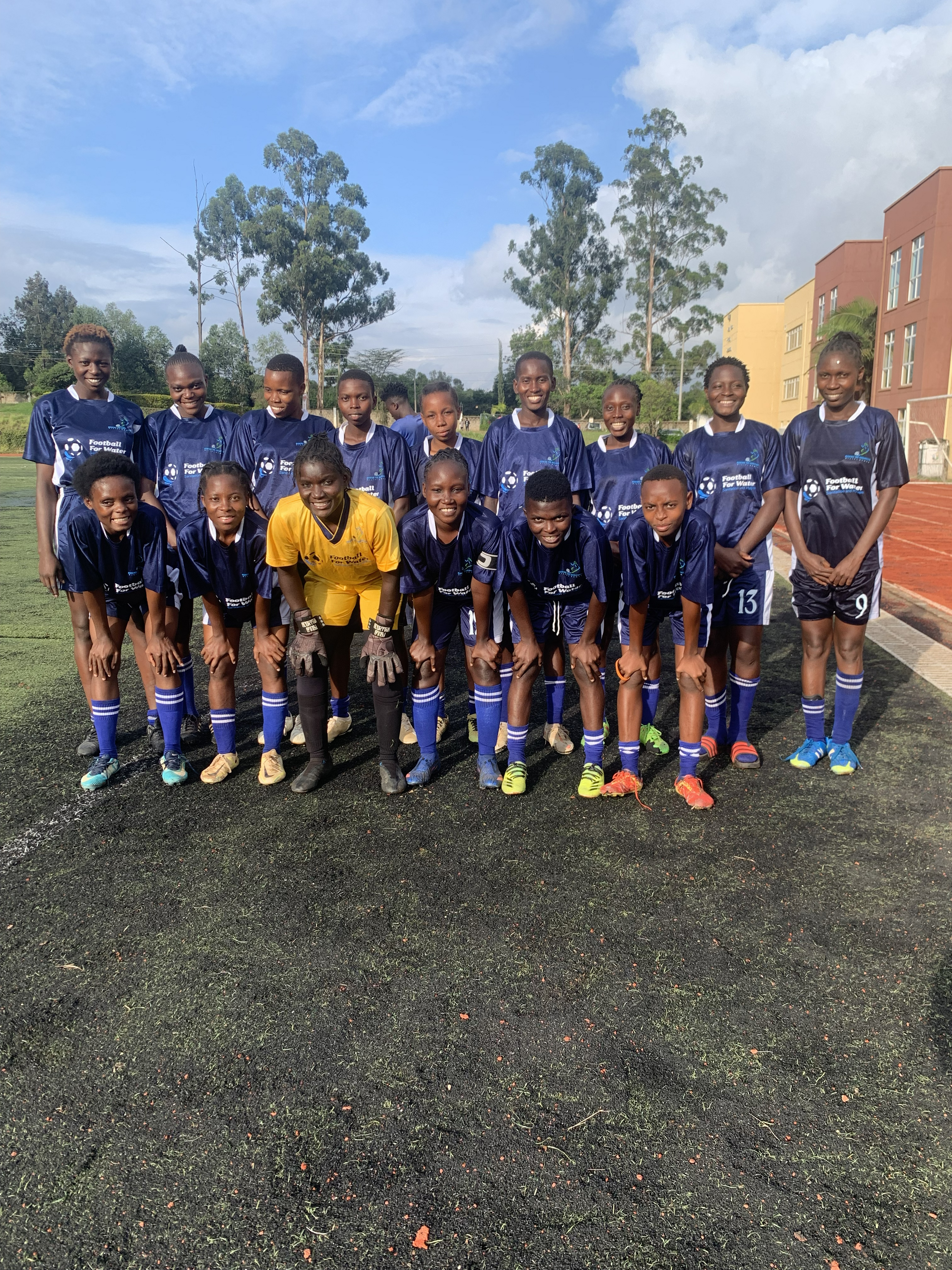 Girls in navy football kits pose with a goalkeeper on an outdoor pitch beside a running track in Kenya.
