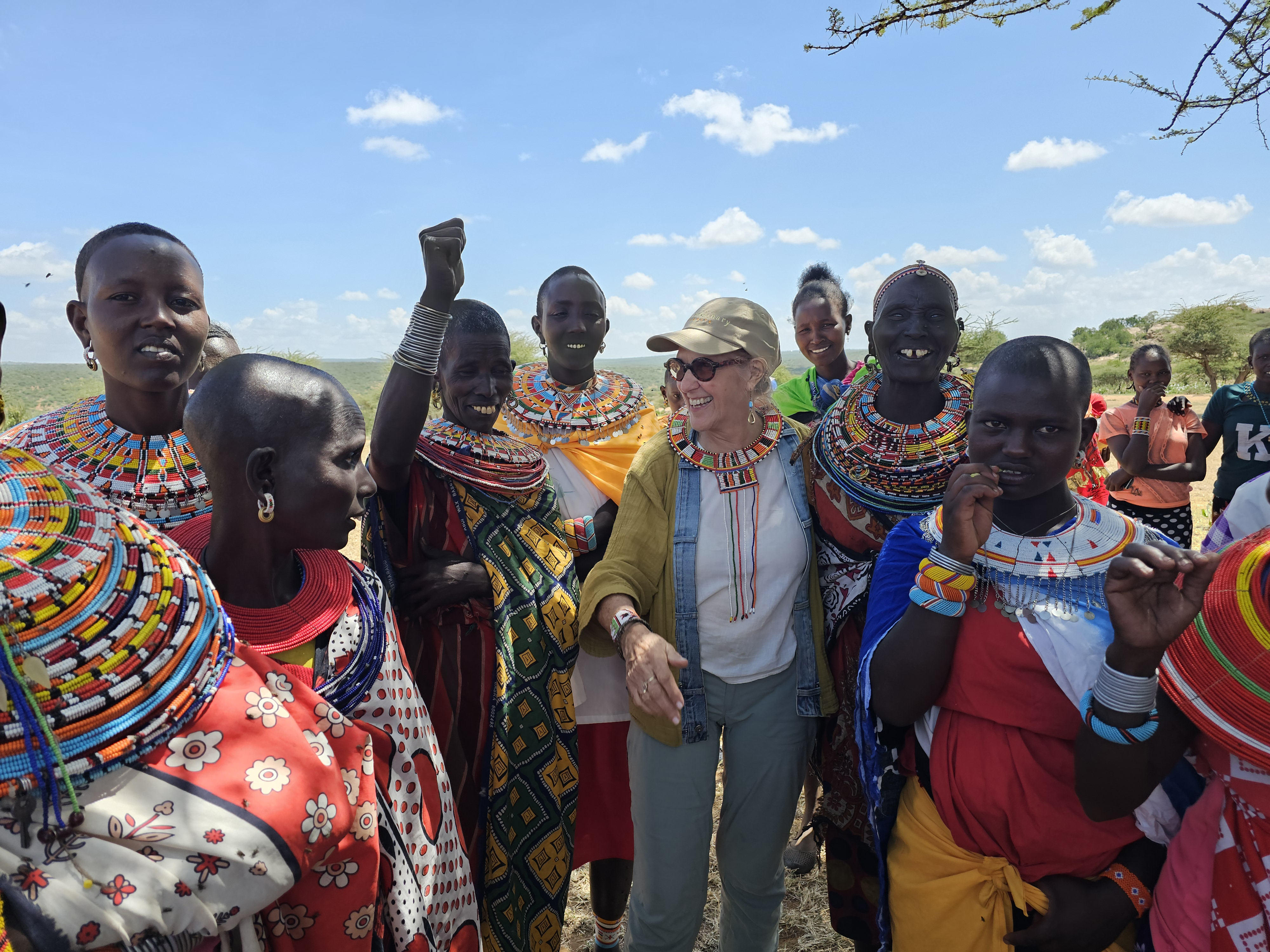A woman in a cap and sunglasses smiles as Samburu women in beaded collars gather around her outdoors in Kenya.