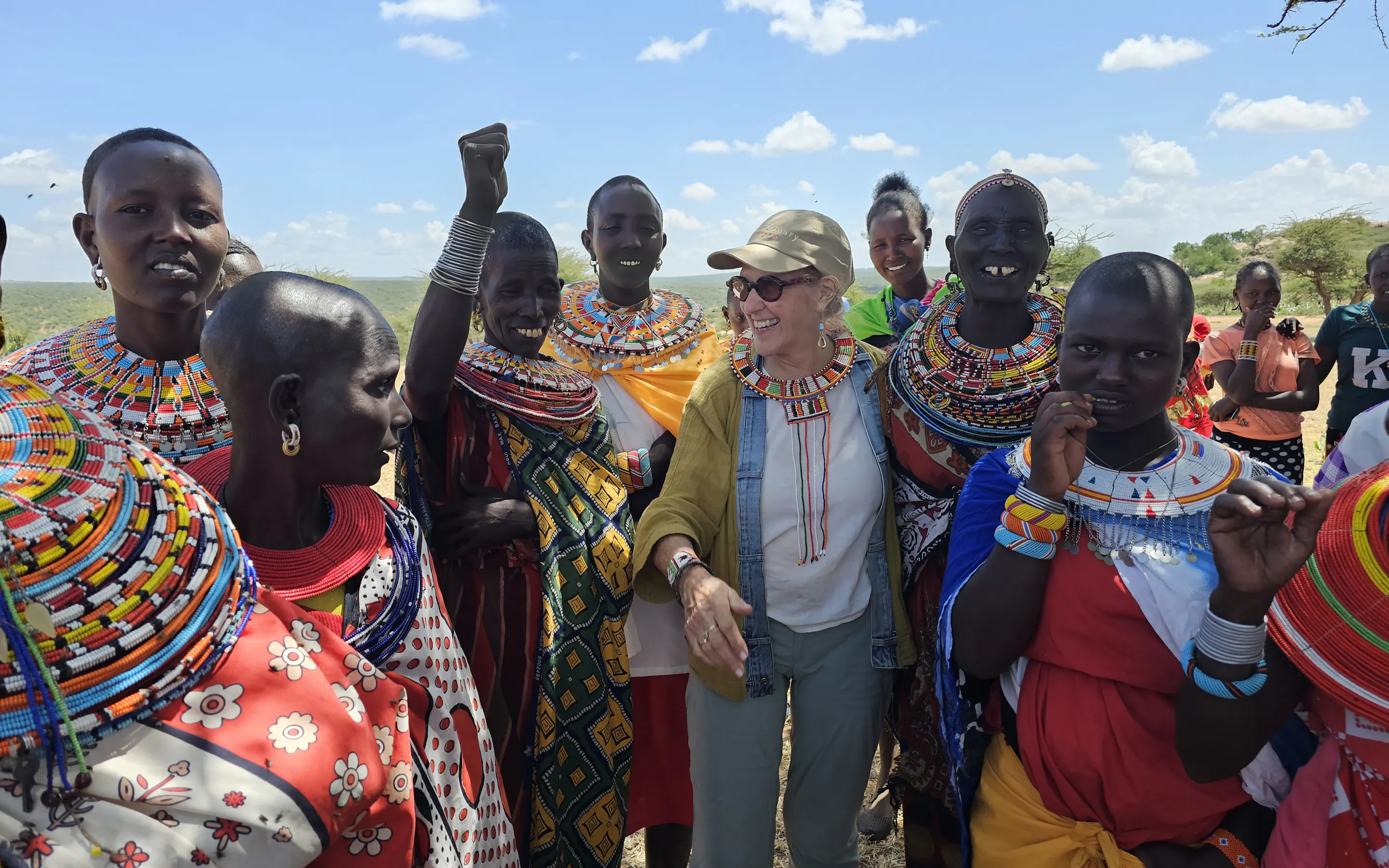 A woman in a cap and sunglasses smiles as Samburu women in beaded collars gather around her outdoors in Kenya.