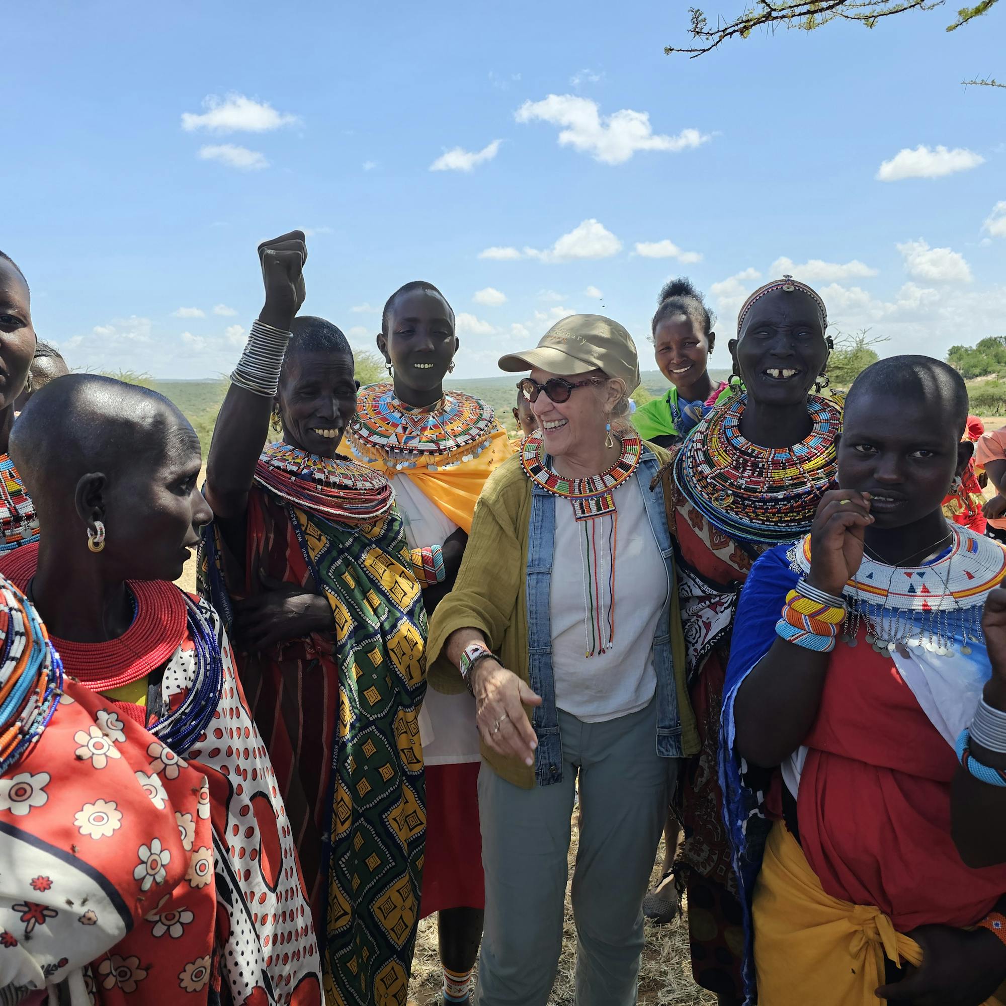 A woman in a cap and sunglasses smiles as Samburu women in beaded collars gather around her outdoors in Kenya.