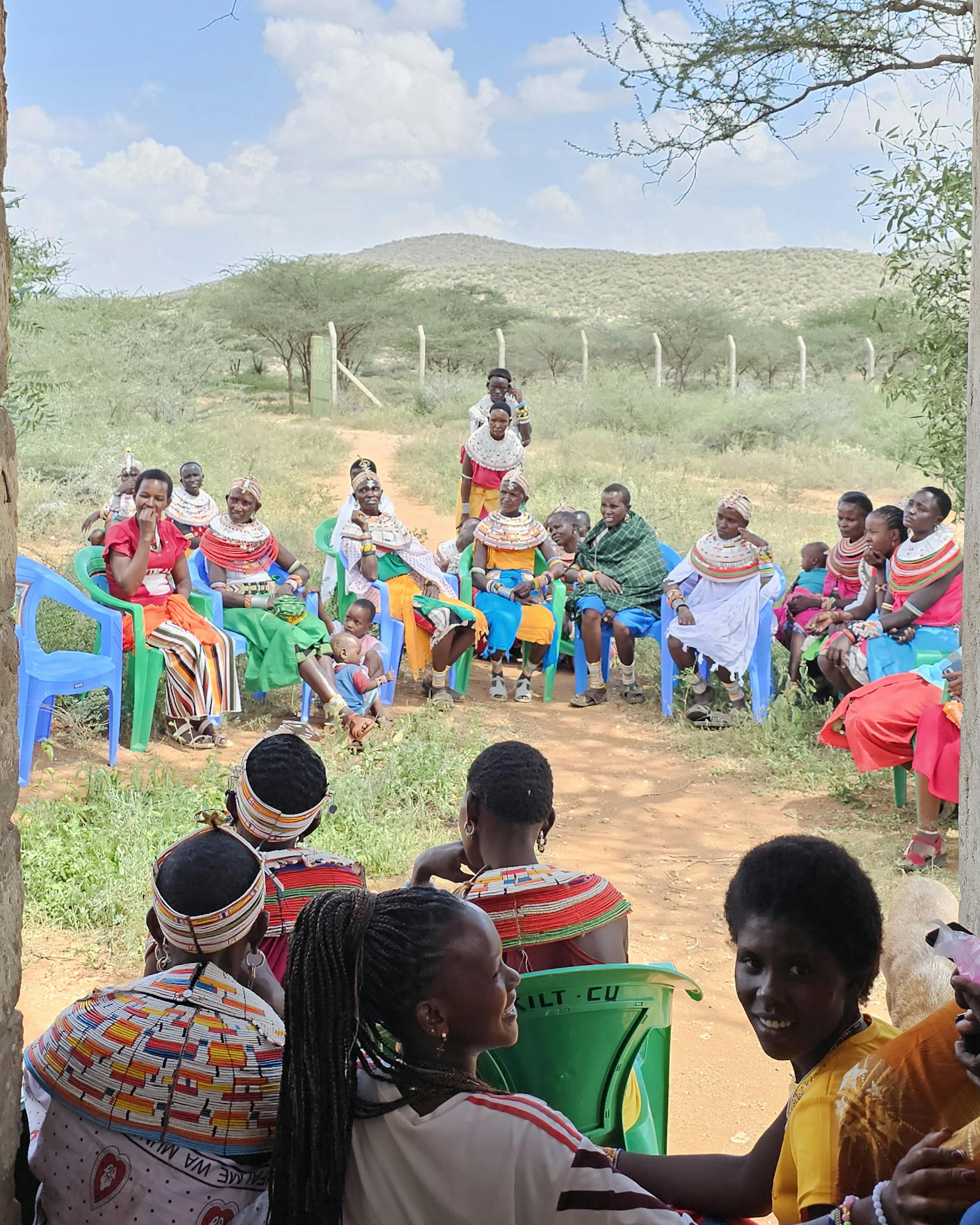 Women in colorful wraps sit in a circle outside as youths watch from a shaded doorway, with hills in rural Kenya.