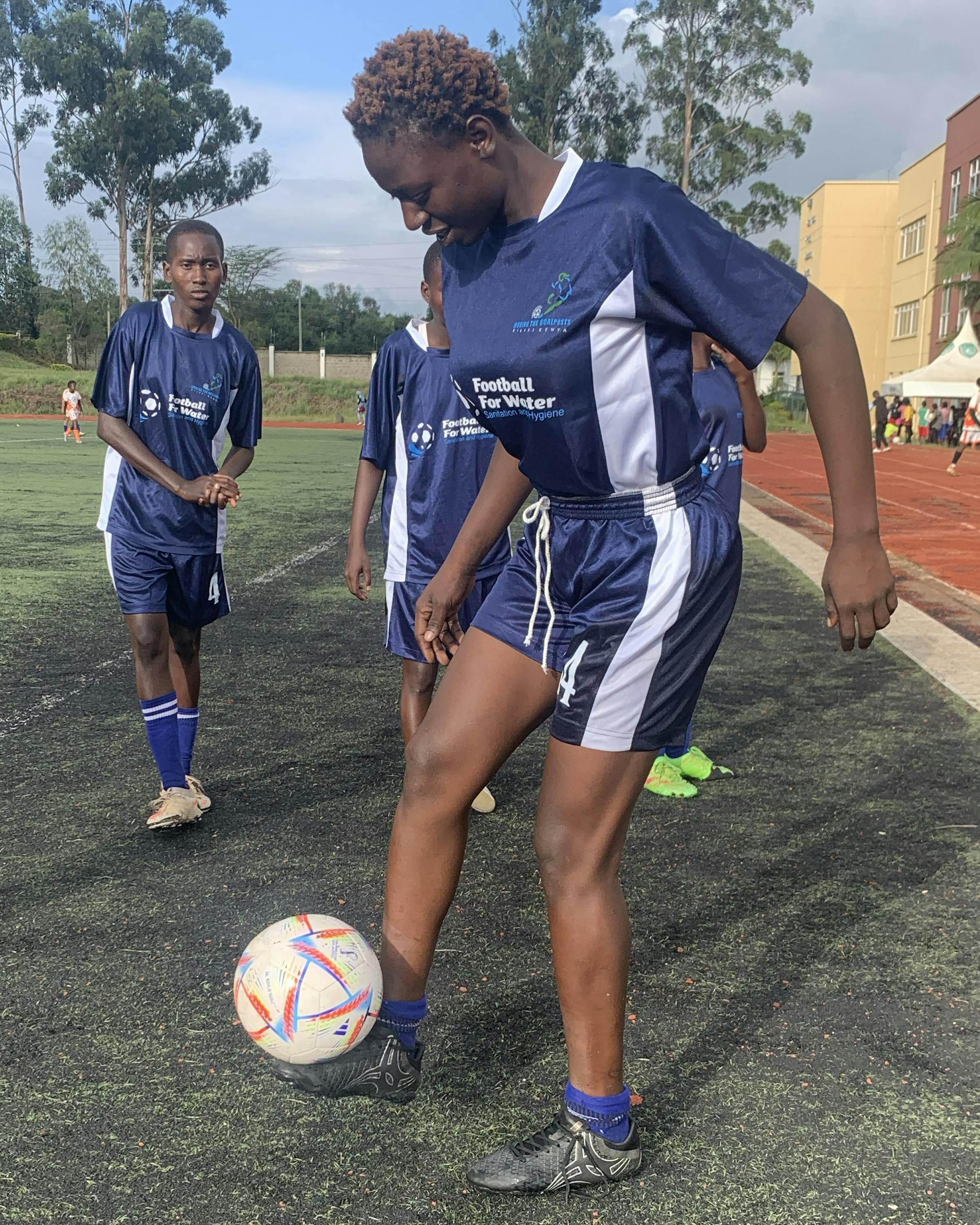 A player in a navy football kit traps a ball on an outdoor pitch beside a running track during a practice in Kenya.