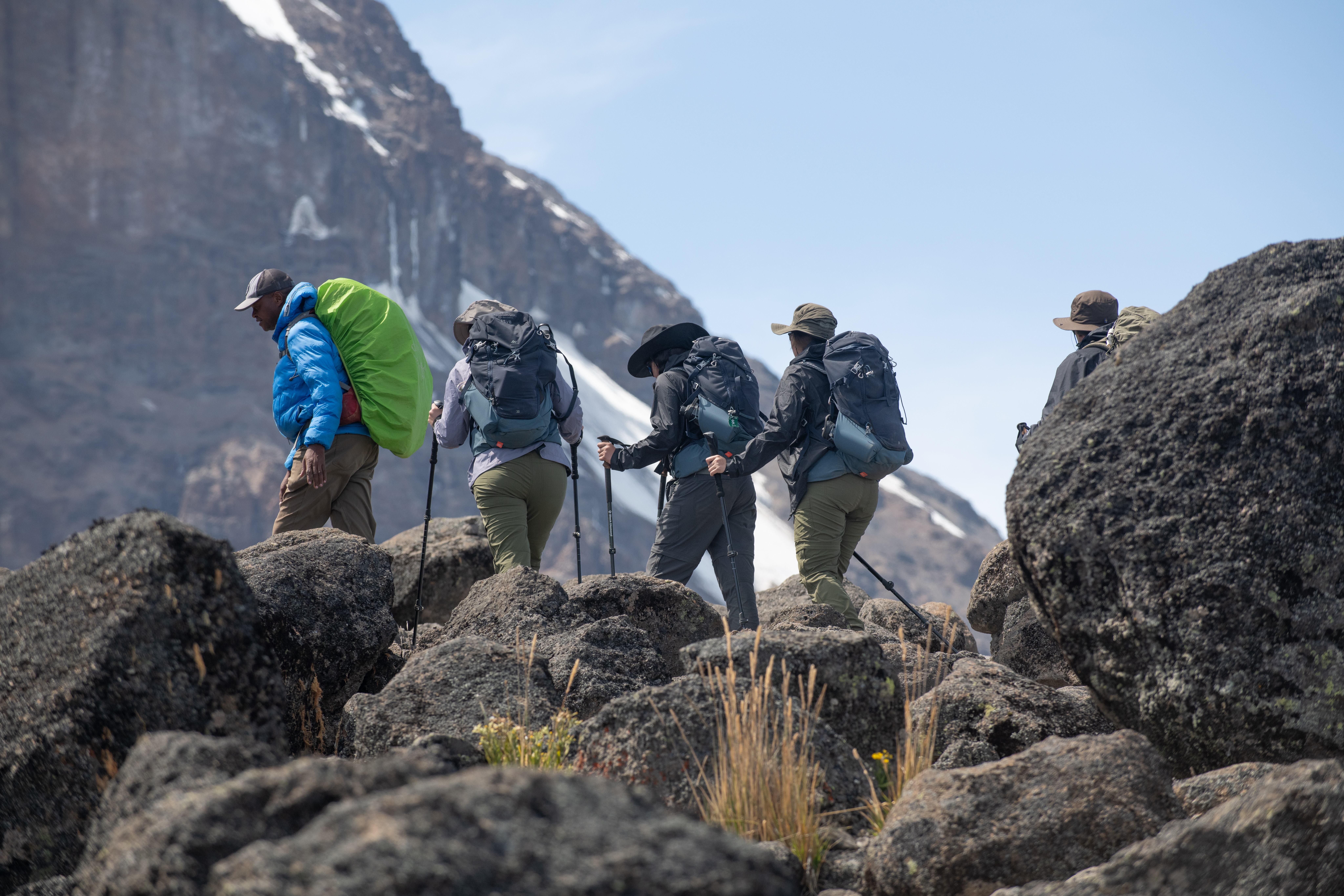 Hikers climb over dark rocks toward a snowy summit ridge, carrying packs high above the clouds in pale morning light.