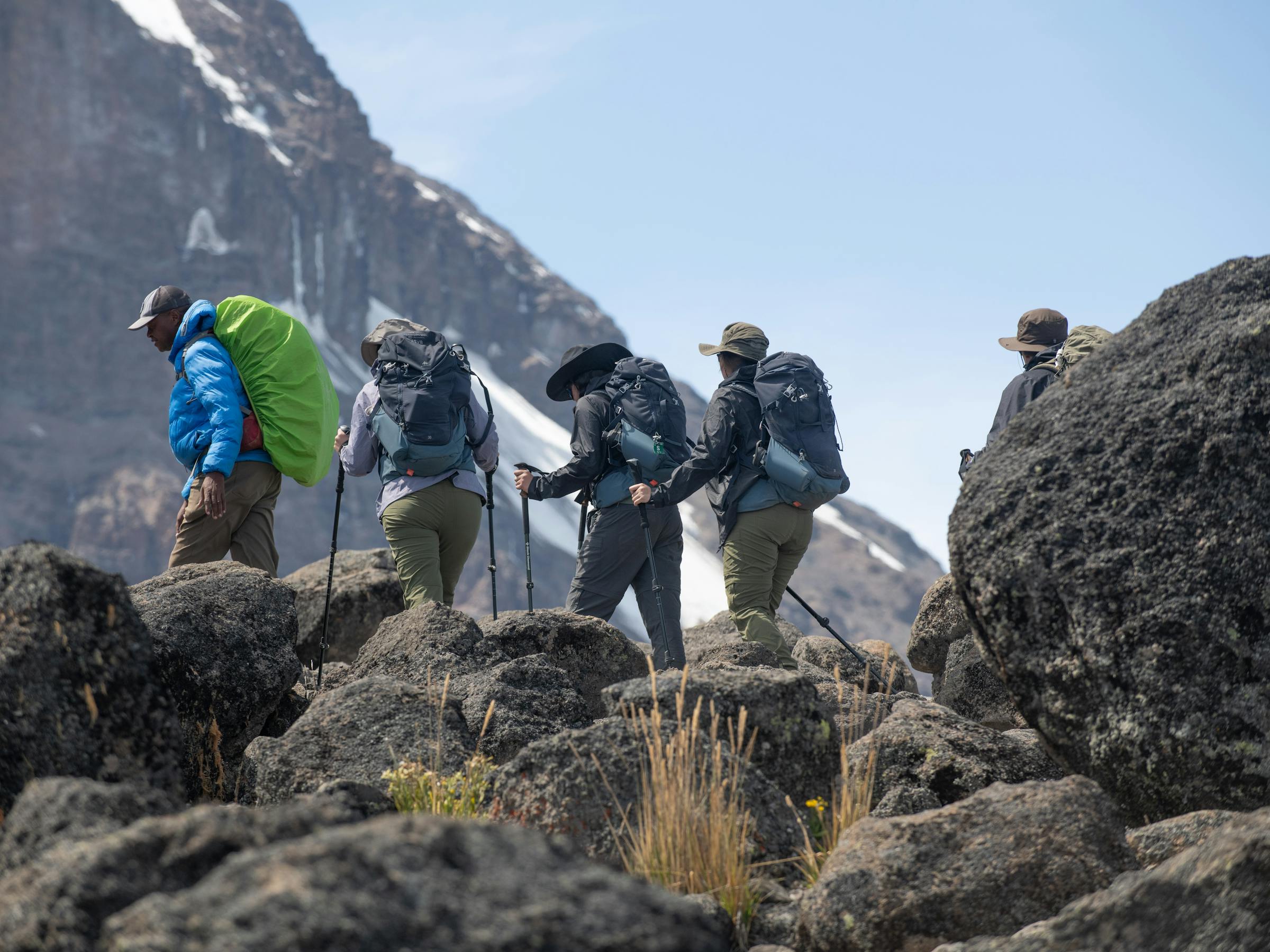 Hikers climb over dark rocks toward a snowy summit ridge, carrying packs high above the clouds in pale morning light.