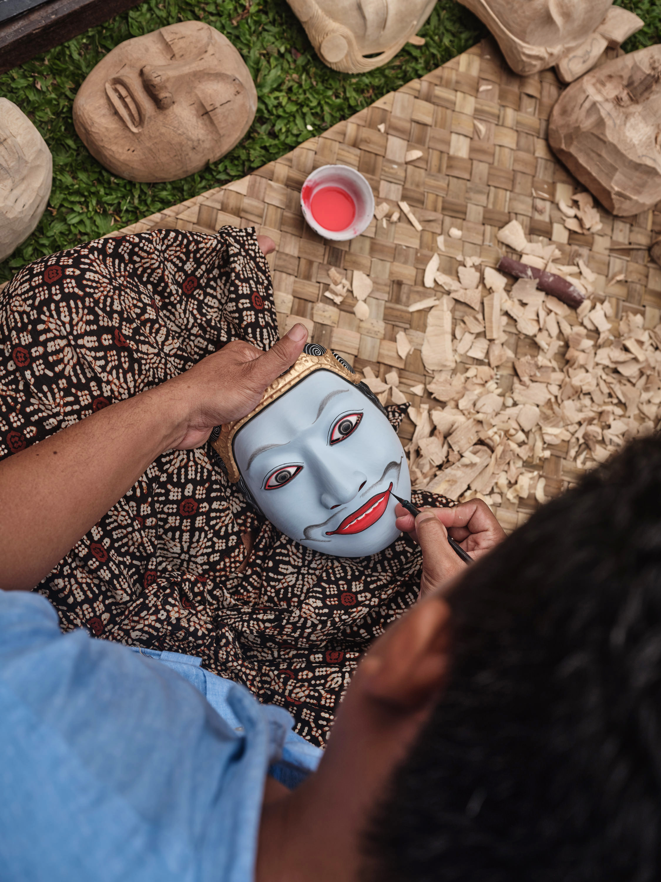 Hands paint a blue mask on a woven mat with art supplies nearby.